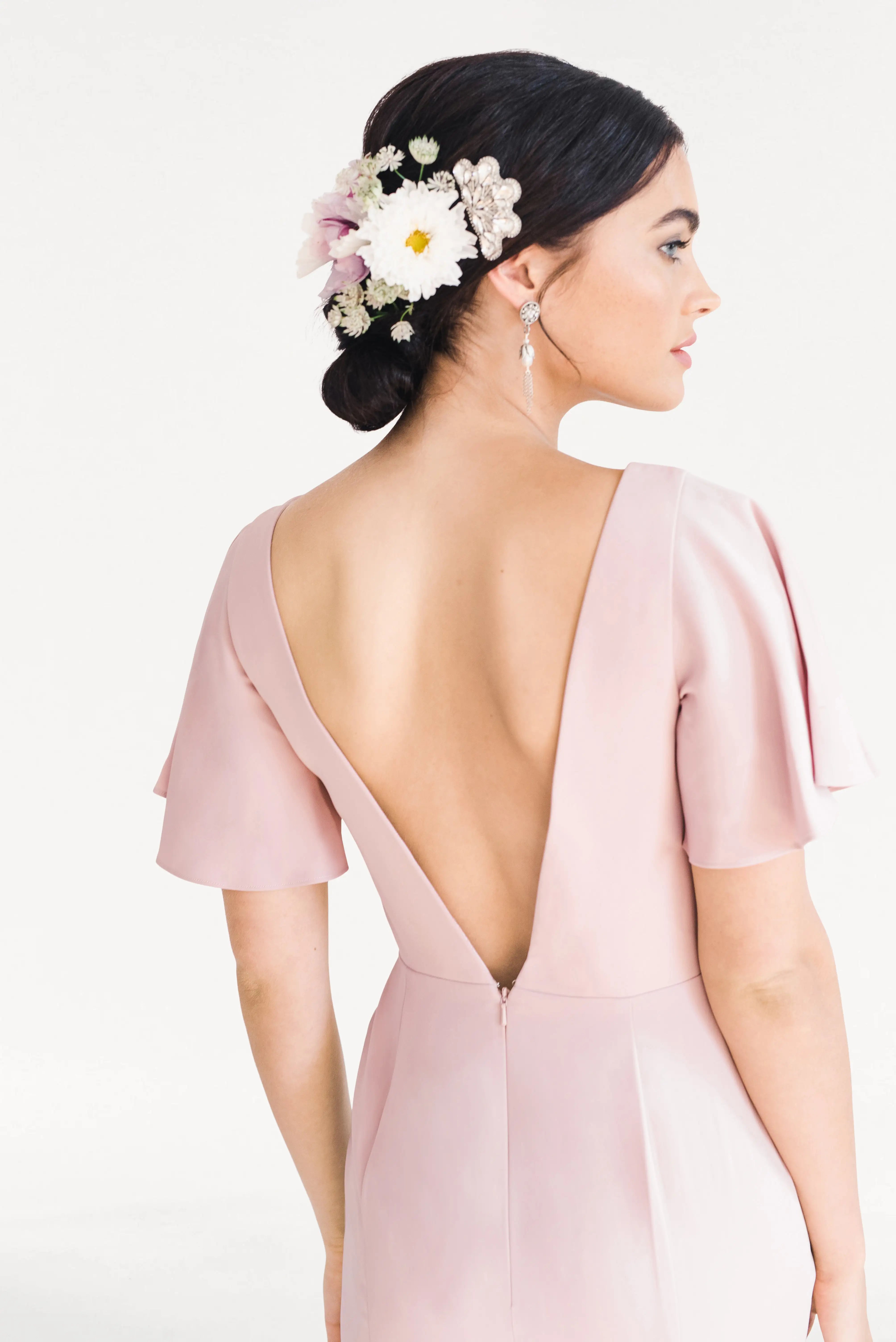 A woman wears the TH&TH Celeste Moss Crepe Bridesmaid Dress in Smoked Blush, posing turned away to reveal a low bun with white and pink flowers, pearl accents, and dangling earrings against a white background.