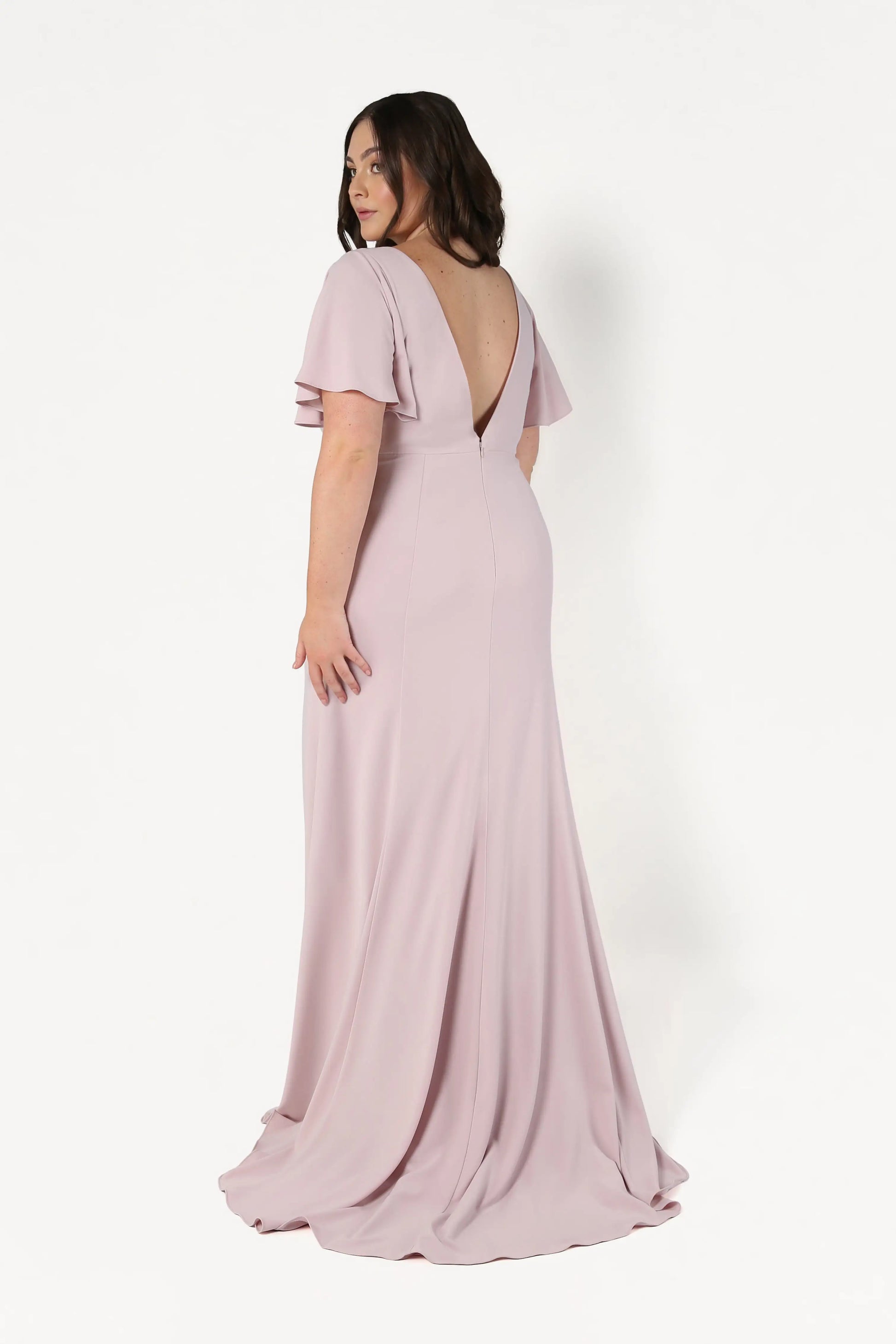 A woman in the TH&TH Celeste Moss Crepe Bridesmaid Dress in Smoked Blush, featuring short flutter sleeves and a deep V-back, stands facing away and looks over her shoulder against a plain white background.