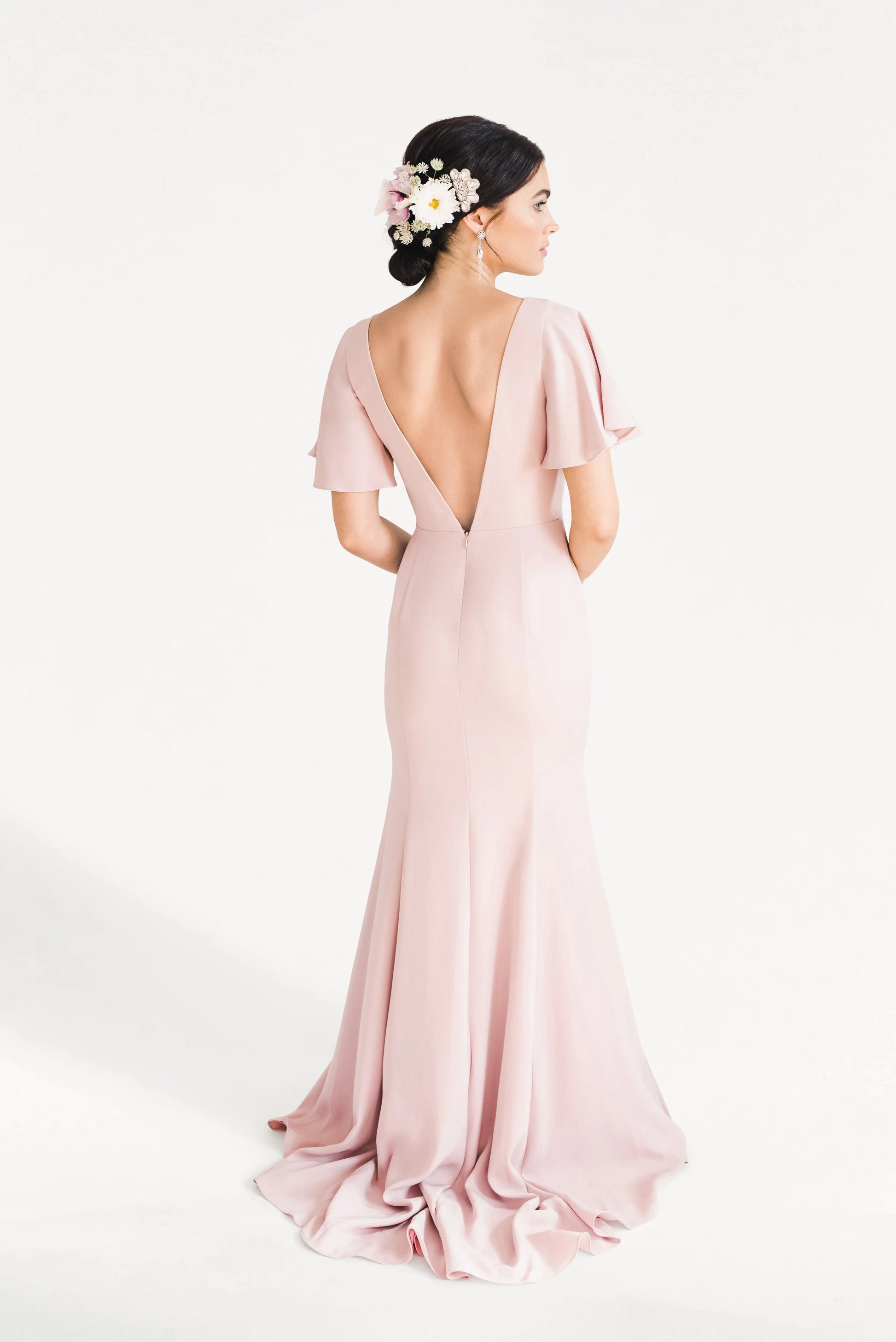 A woman with dark hair styled in an updo adorned with pink and white flowers wears the TH&TH Celeste Moss Crepe Bridesmaid Dress in Smoked Blush, featuring flutter sleeves and a deep V-back, standing against a plain white background.