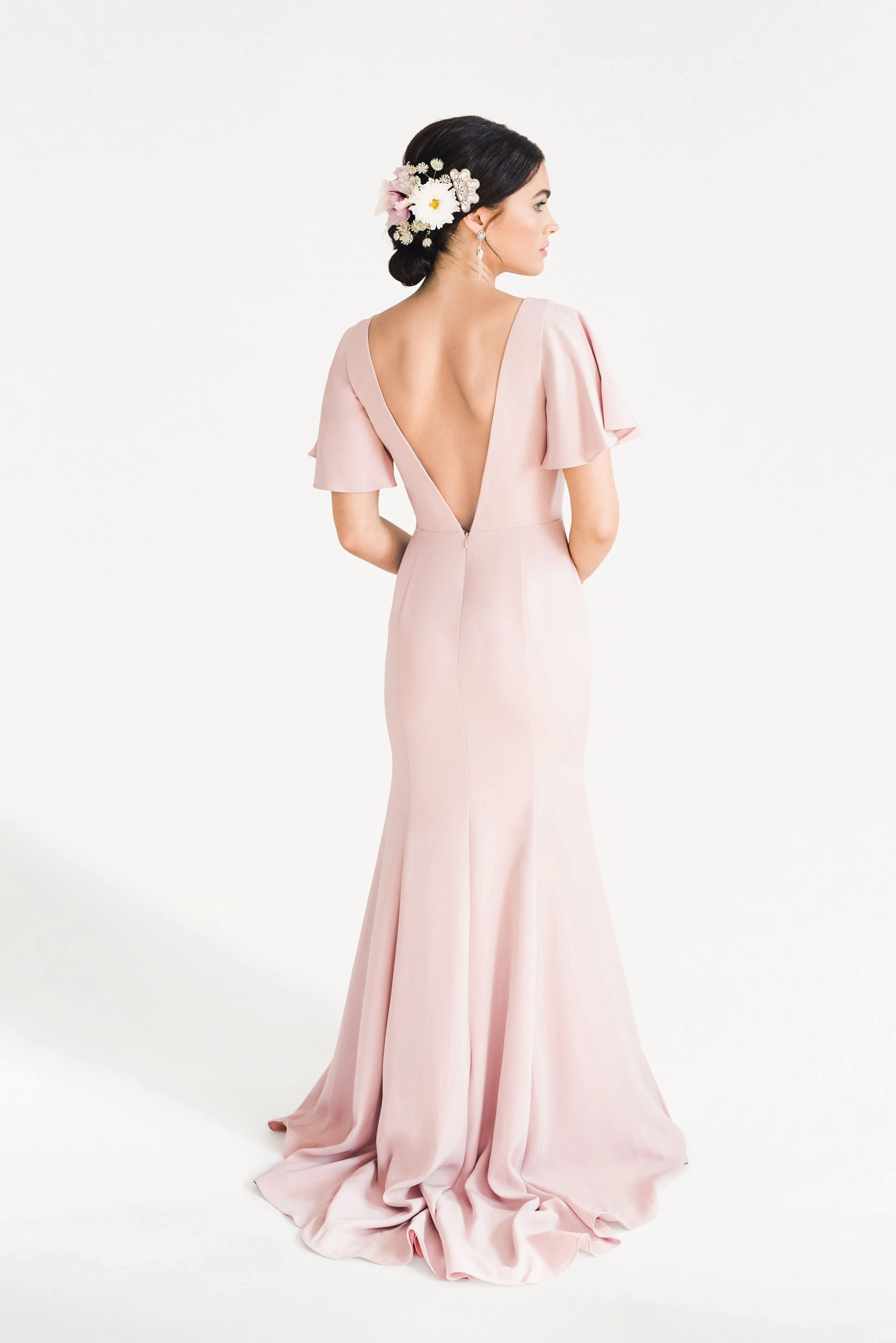 A woman with dark hair styled in an updo adorned with pink and white flowers wears the TH&TH Celeste Moss Crepe Bridesmaid Dress in Smoked Blush, featuring flutter sleeves and a deep V-back, standing against a plain white background.