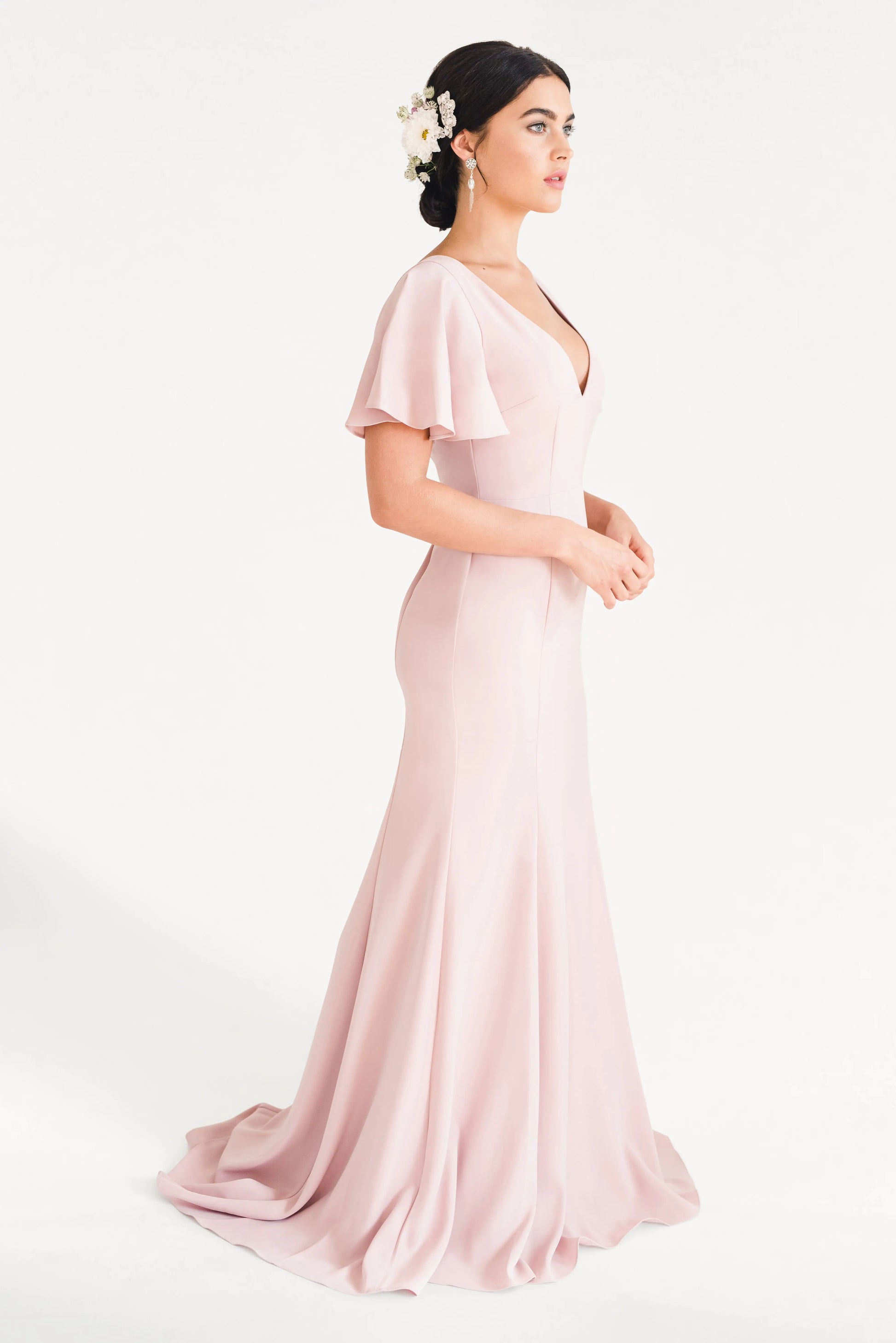 A woman stands in profile wearing the TH&TH Celeste Moss Crepe Bridesmaid Dress in Smoked Blush, a floor-length gown with flutter sleeves. Her low bun, adorned with white flowers, complements the elegant look against a plain white background.