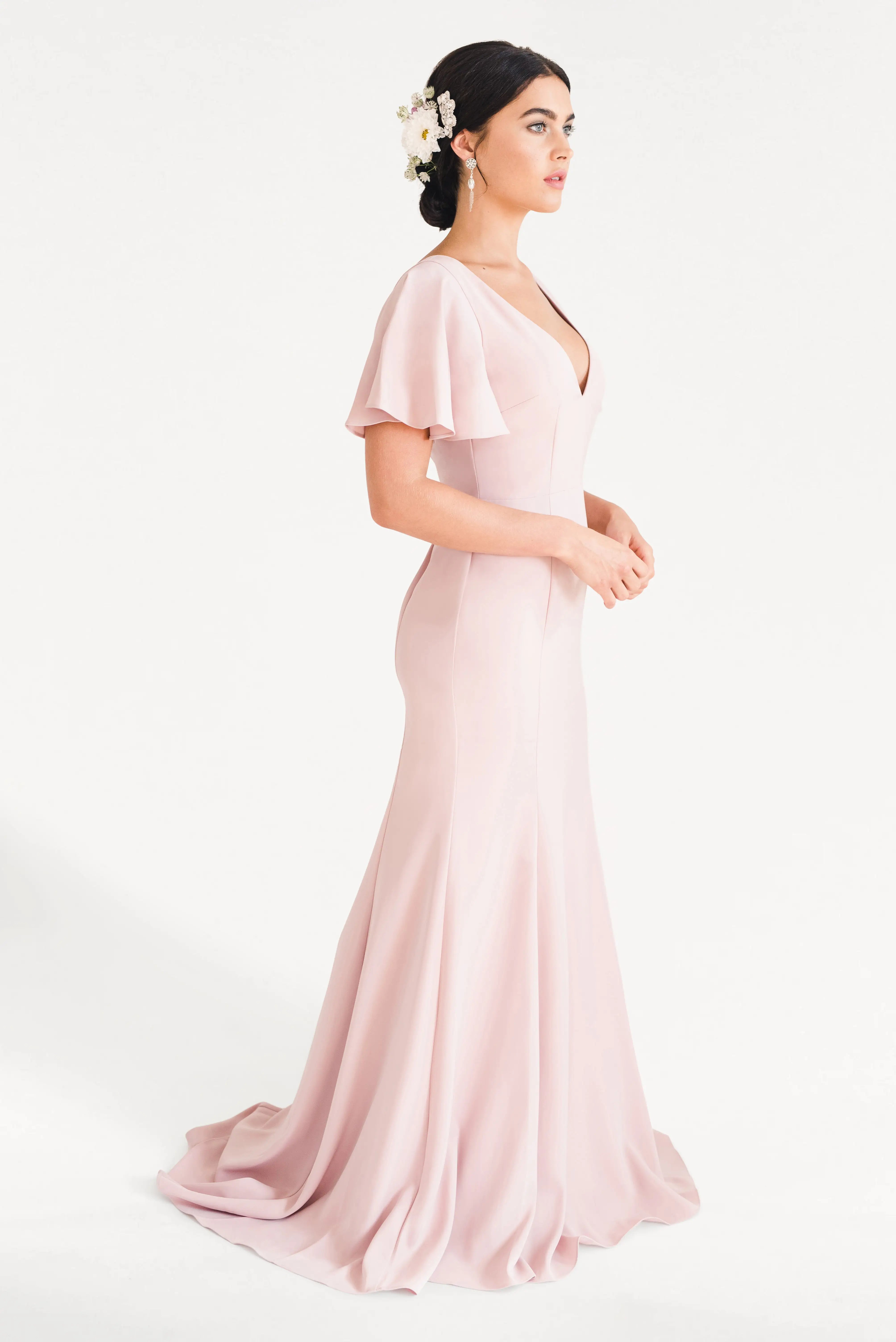 A woman stands in profile wearing the TH&TH Celeste Moss Crepe Bridesmaid Dress in Smoked Blush, a floor-length gown with flutter sleeves. Her low bun, adorned with white flowers, complements the elegant look against a plain white background.