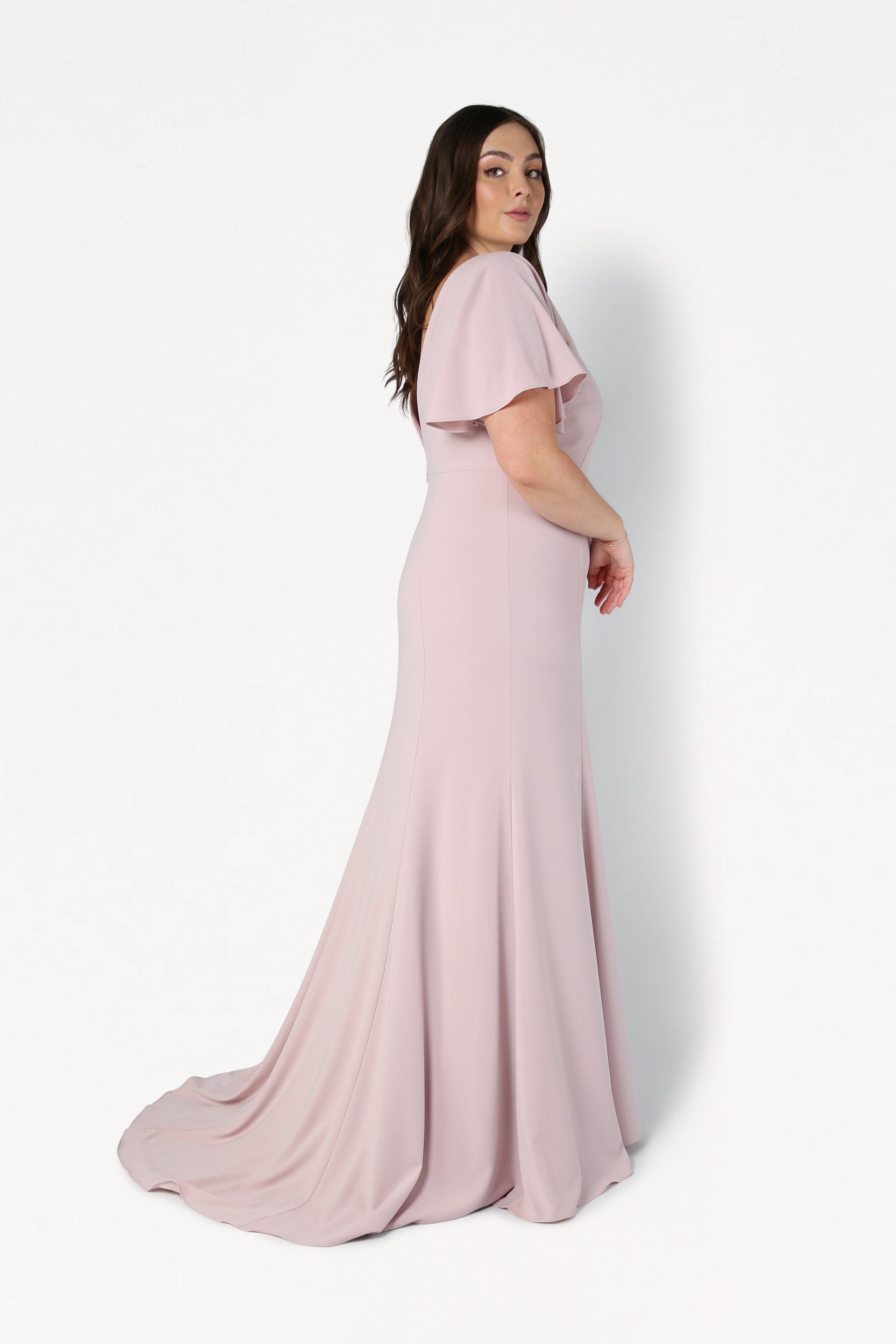 A woman with long brown hair stands sideways against a white background, wearing the TH&TH Celeste Moss Crepe Bridesmaid Dress in Smoked Blush. This floor-length gown features short flutter sleeves, a fitted bodice, and a gentle train for an elegant look.