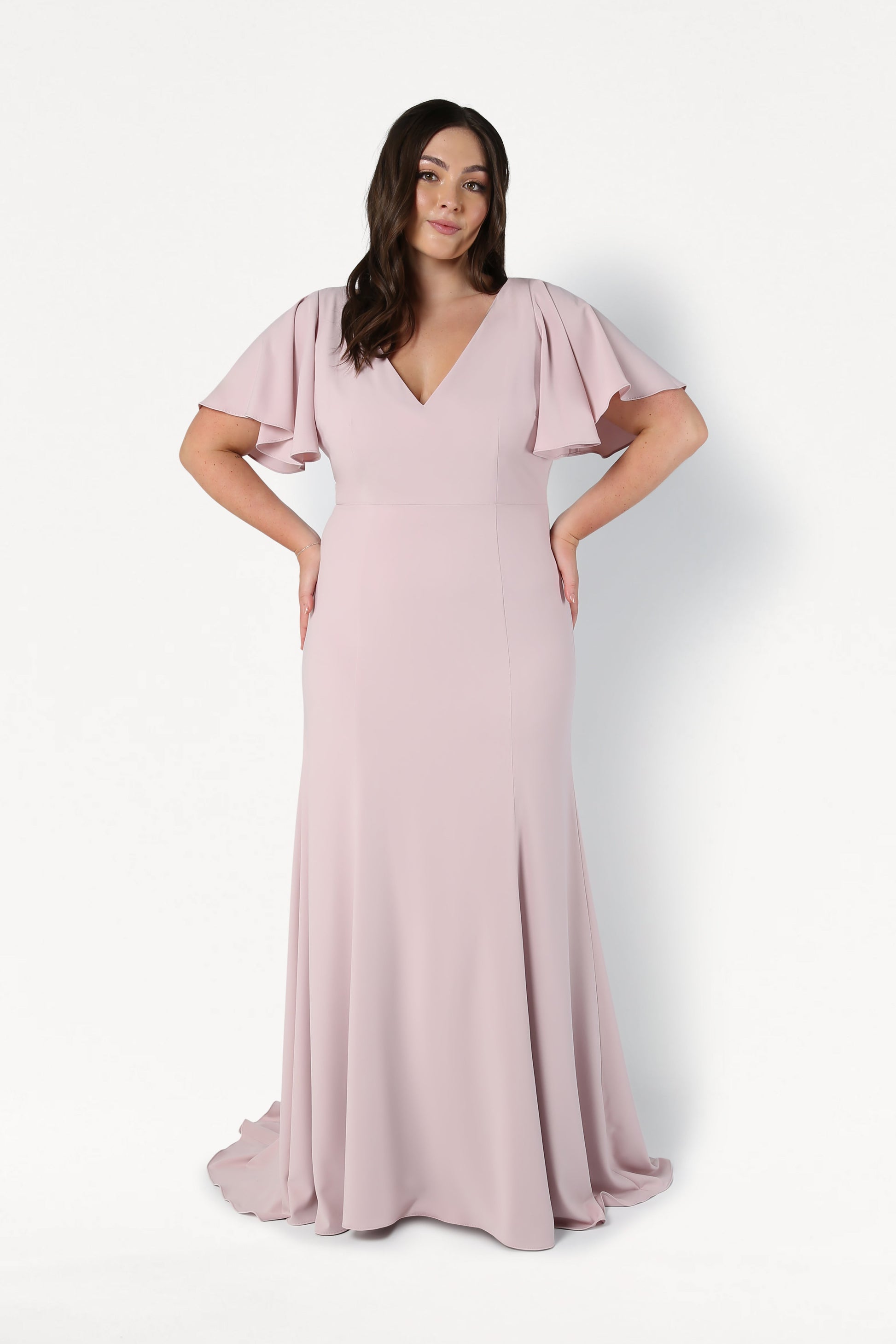 A woman with long dark hair stands against a white background wearing the TH&TH Celeste Moss Crepe Bridesmaid Dress in Smoked Blush, featuring a floor-length skirt, flutter sleeves, and a V-neckline. Her hands rest on her hips.