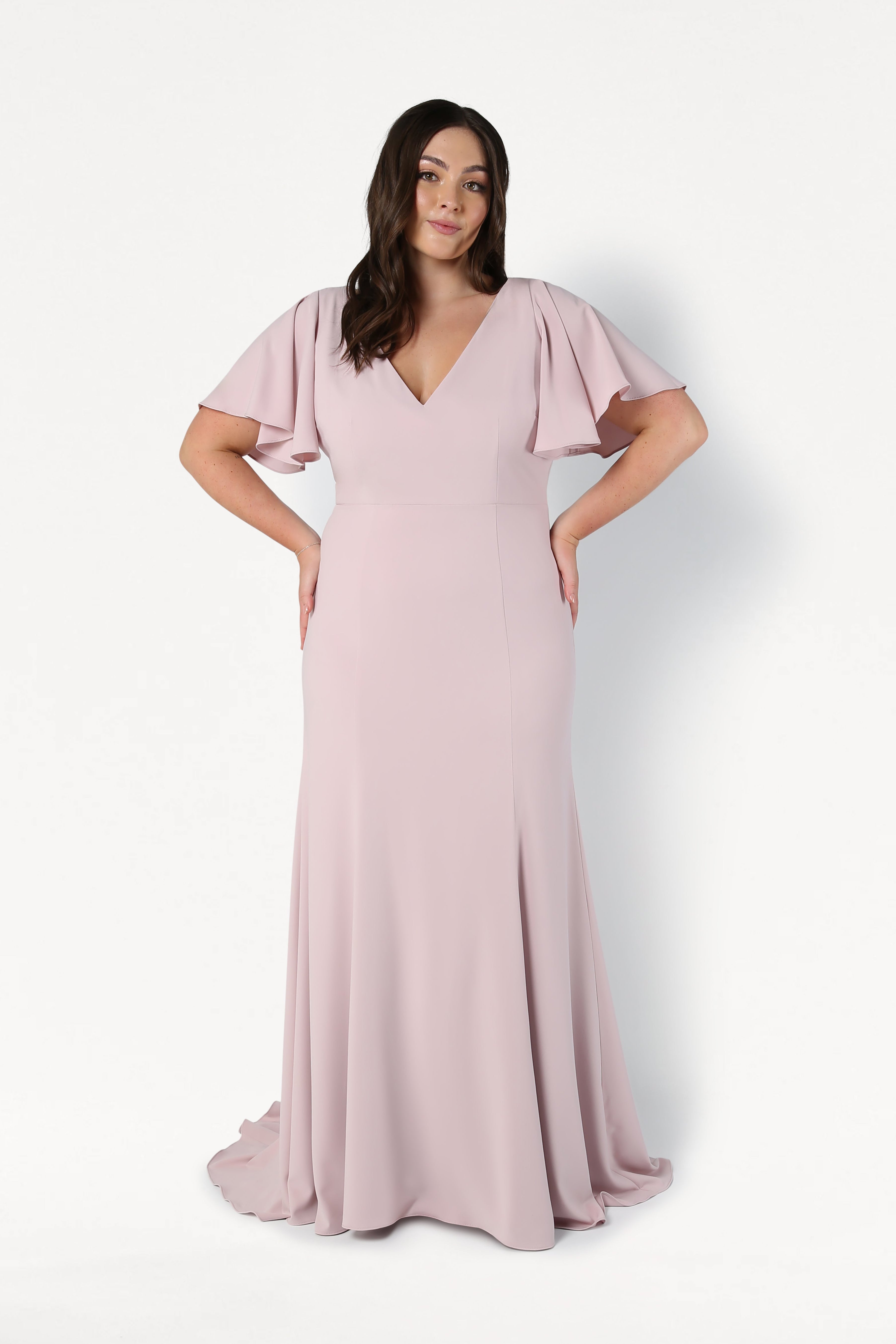 A woman with long dark hair stands against a white background wearing the TH&TH Celeste Moss Crepe Bridesmaid Dress in Smoked Blush, featuring a floor-length skirt, flutter sleeves, and a V-neckline. Her hands rest on her hips.