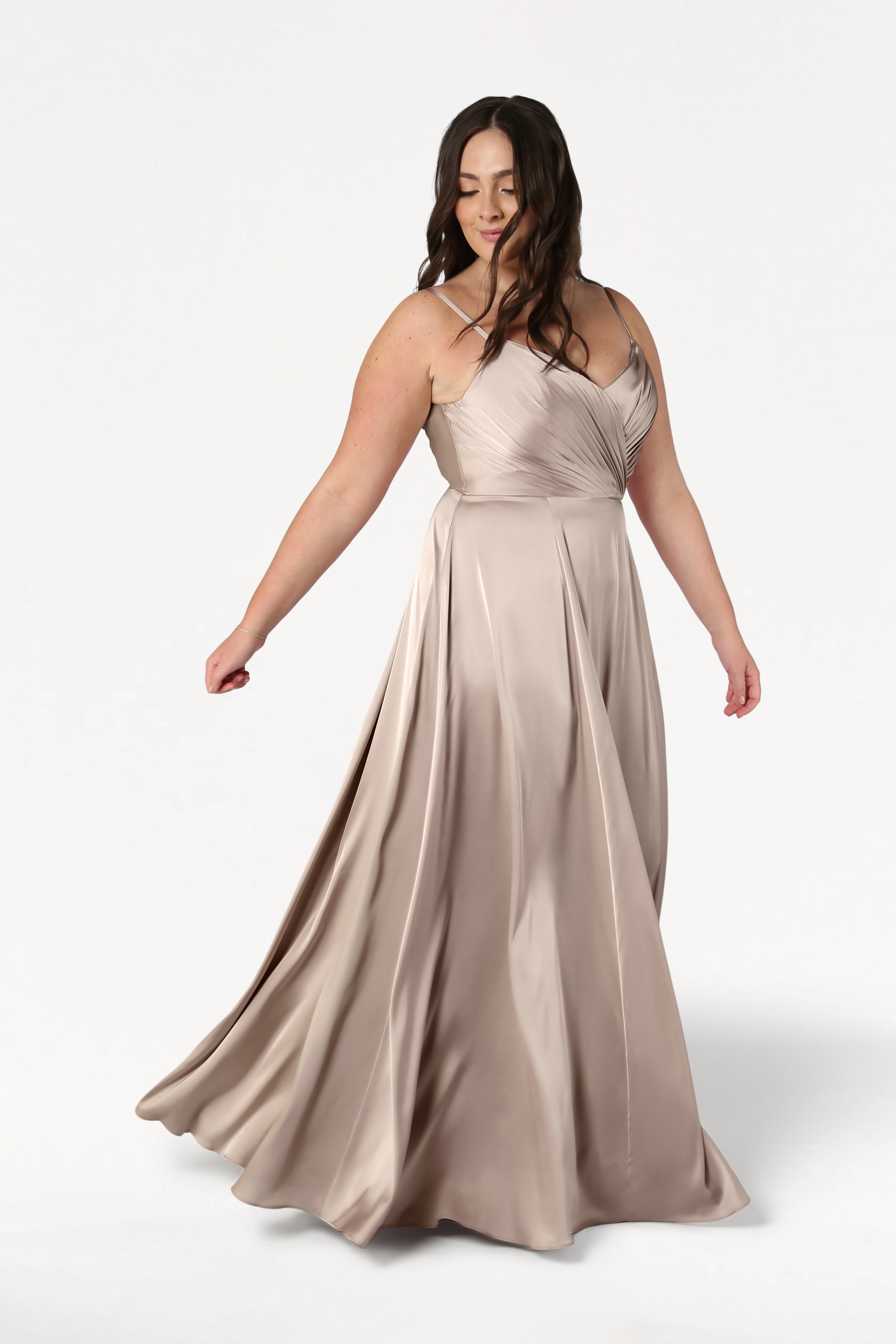 A woman twirls gracefully in the TH&TH Isla Bridesmaid Dress in Champagne Sky Satin, featuring a long flowing skirt, pleated bodice, and spaghetti straps, set against a plain white background.