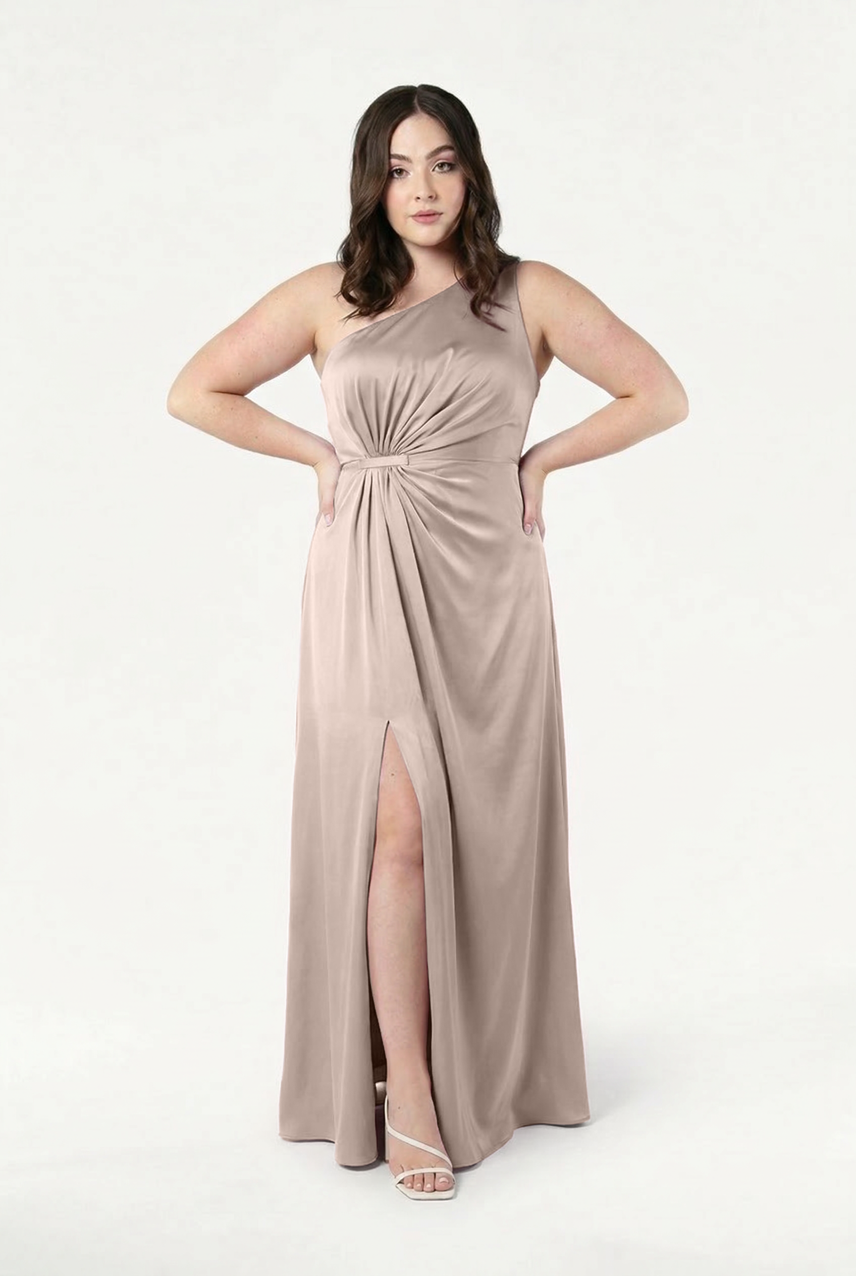 A woman faces forward in the Greta Bridesmaid Dress in Champagne Sky Satin by TH&TH, featuring a waist twist detail and front slit. She has wavy brown hair, white heeled sandals, and stands against a plain white background.