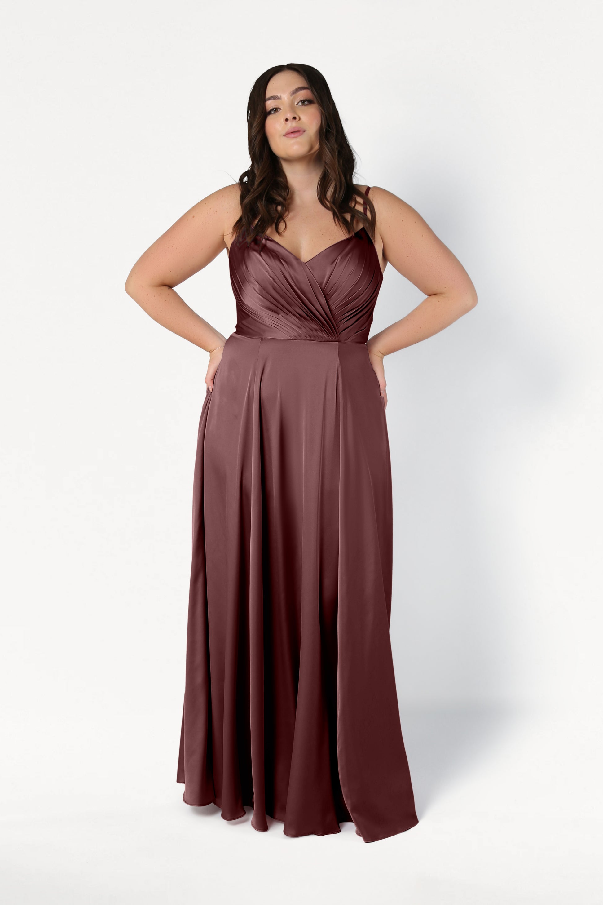 A woman wearing the TH&TH Isla Bridesmaid Dress in chocolate brown satin—a sleeveless, floor-length gown with a pleated bodice—stands against a plain white background.