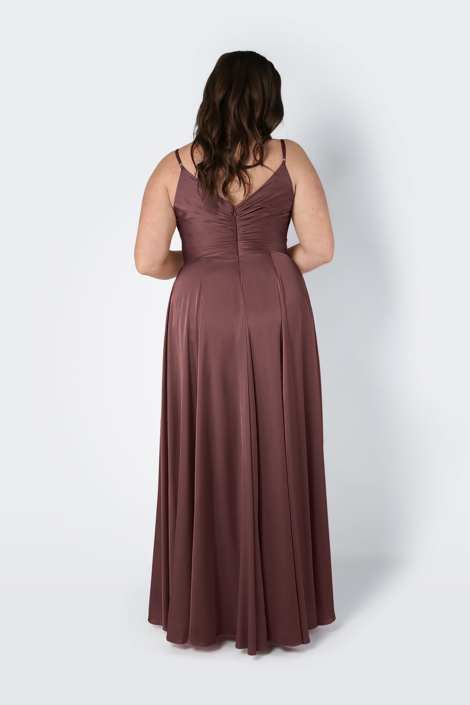 A woman with wavy brown hair stands with her back to the camera, wearing the Isla Bridesmaid Dress in chocolate brown satin by TH&TH—a sleeveless, floor-length dress with thin straps and a pleated bodice. The background is plain and light-colored.