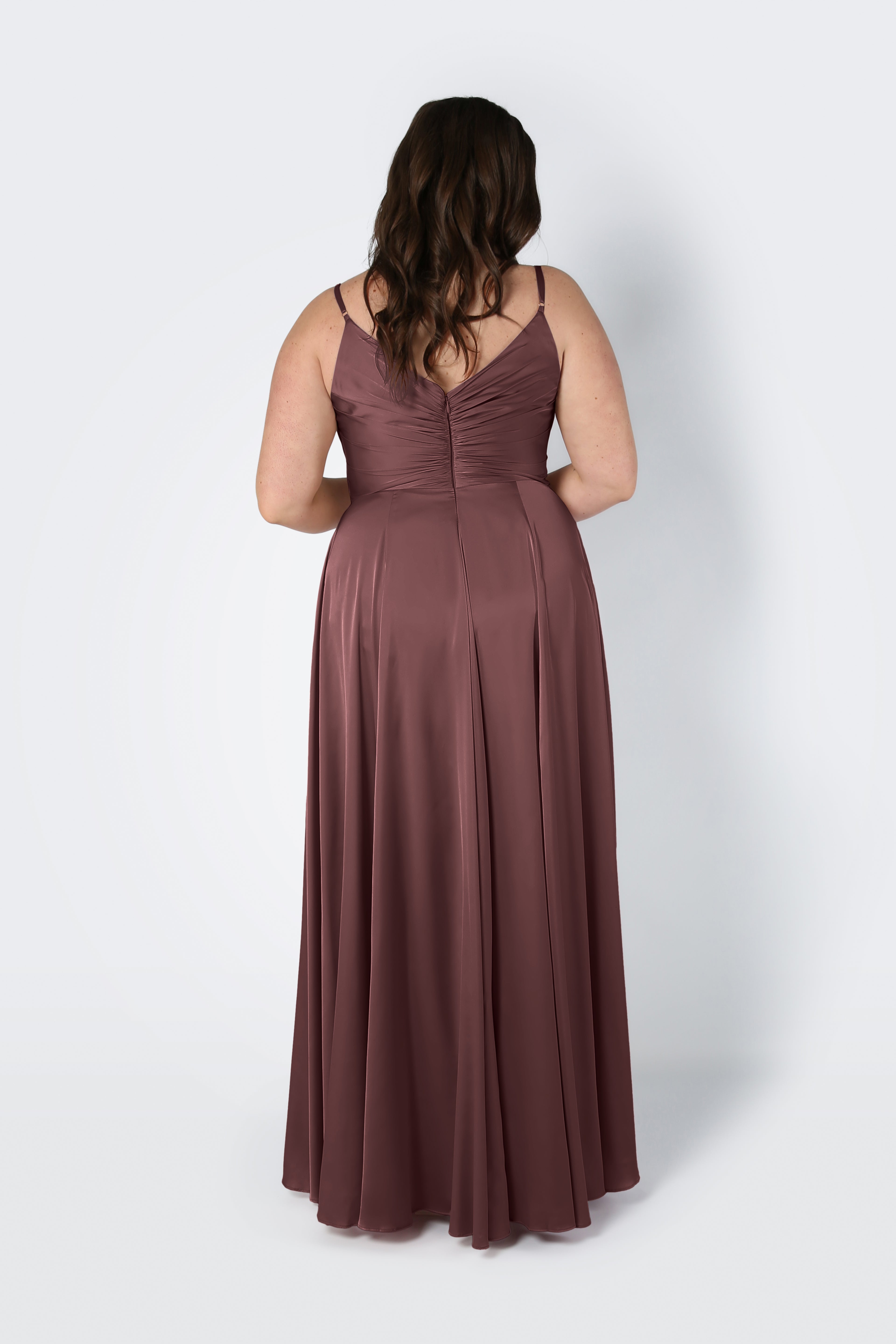 A woman with wavy brown hair stands with her back to the camera, wearing the Isla Bridesmaid Dress in chocolate brown satin by TH&TH—a sleeveless, floor-length dress with thin straps and a pleated bodice. The background is plain and light-colored.
