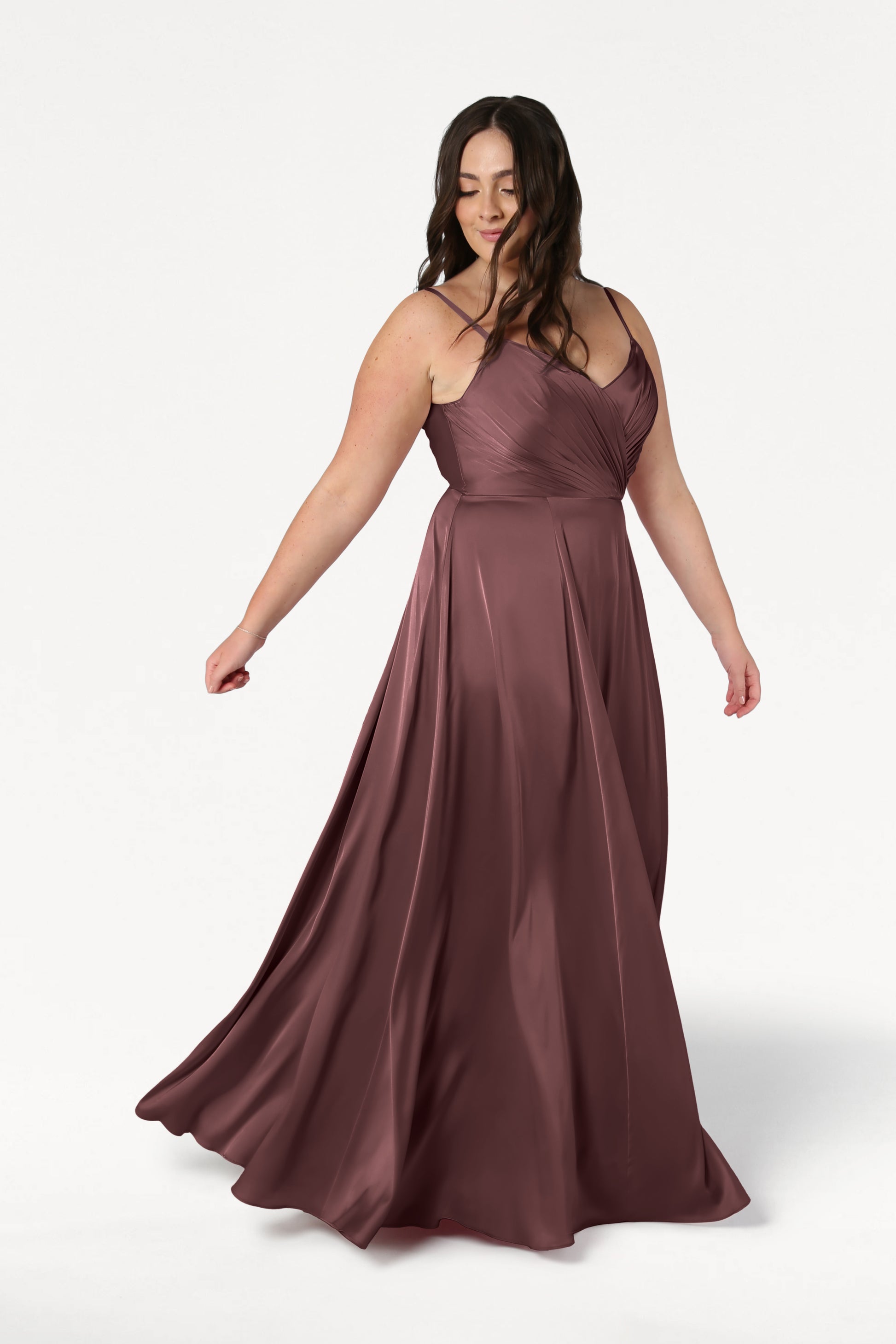 A woman in a chocolate brown satin Isla Bridesmaid Dress by TH&TH twirls gracefully against a white background, showcasing the pleated bodice and flowing skirt.