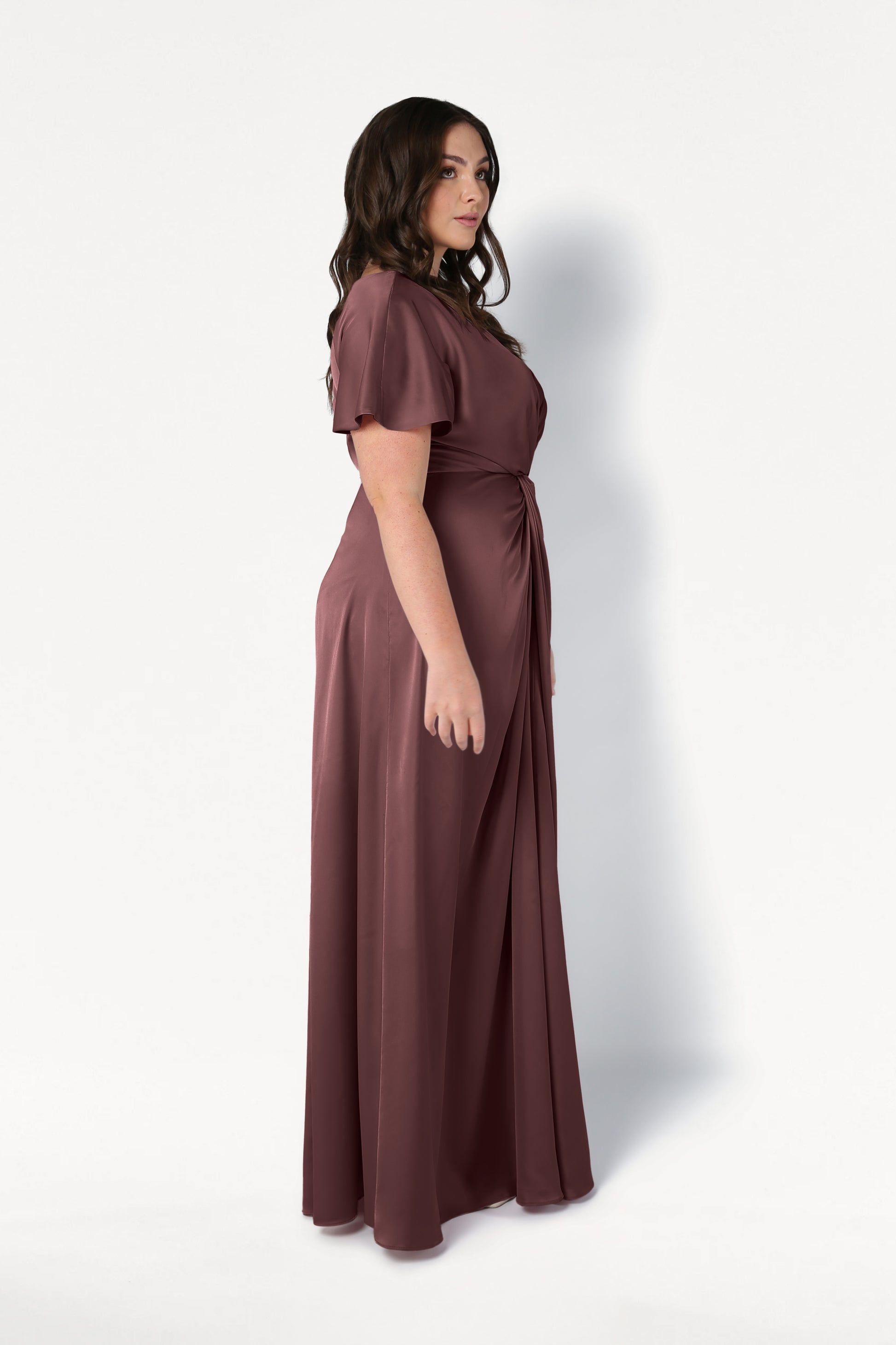 A woman with wavy brown hair stands in profile, modeling the floor-length Camilla Bridesmaid Dress in chocolate brown satin by TH&TH, highlighting its gathered waist detail and size-inclusive design against a plain white background.