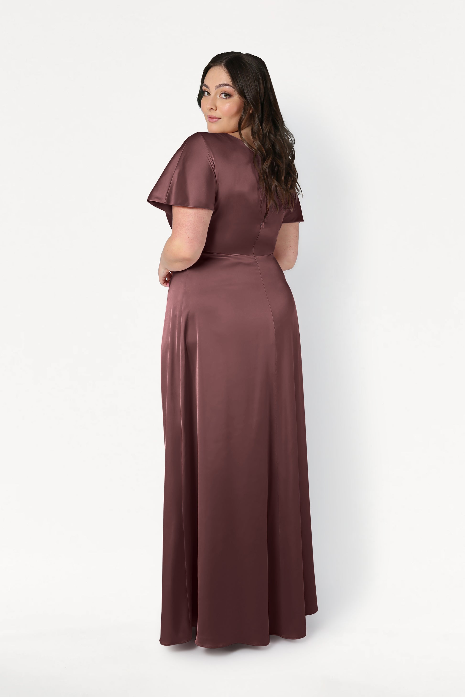A woman with long brown hair stands facing backward, looking over her shoulder. She wears the TH&TH Camilla Bridesmaid Dress in chocolate brown satin against a plain white background.