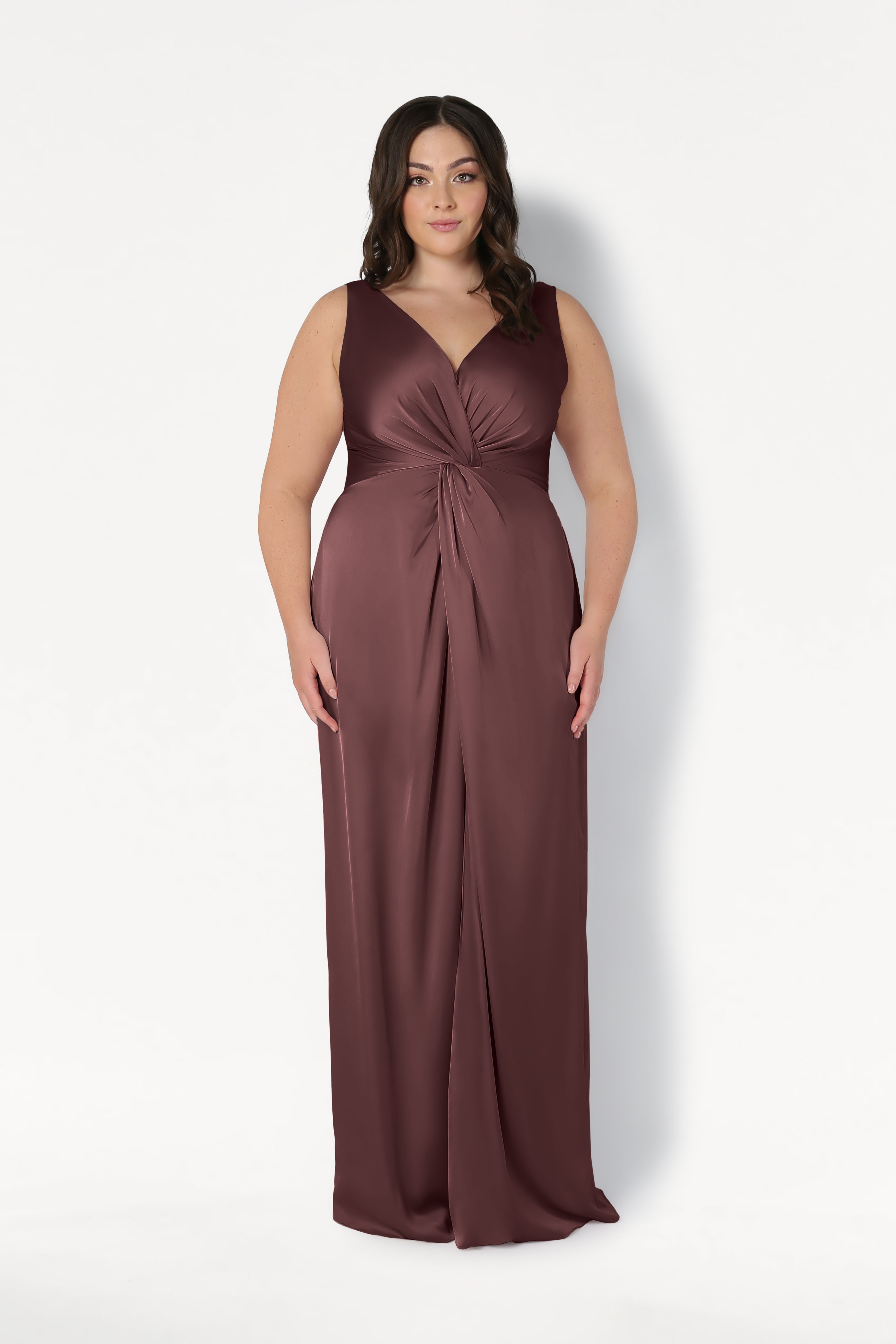 A woman with long dark hair models the Carmen Bridesmaid Dress in Chocolate Brown Satin by TH&TH—a sleeveless, floor-length gown with a V-neckline and gathered knot waist—perfect for modern bridal parties. She stands against a plain white background.