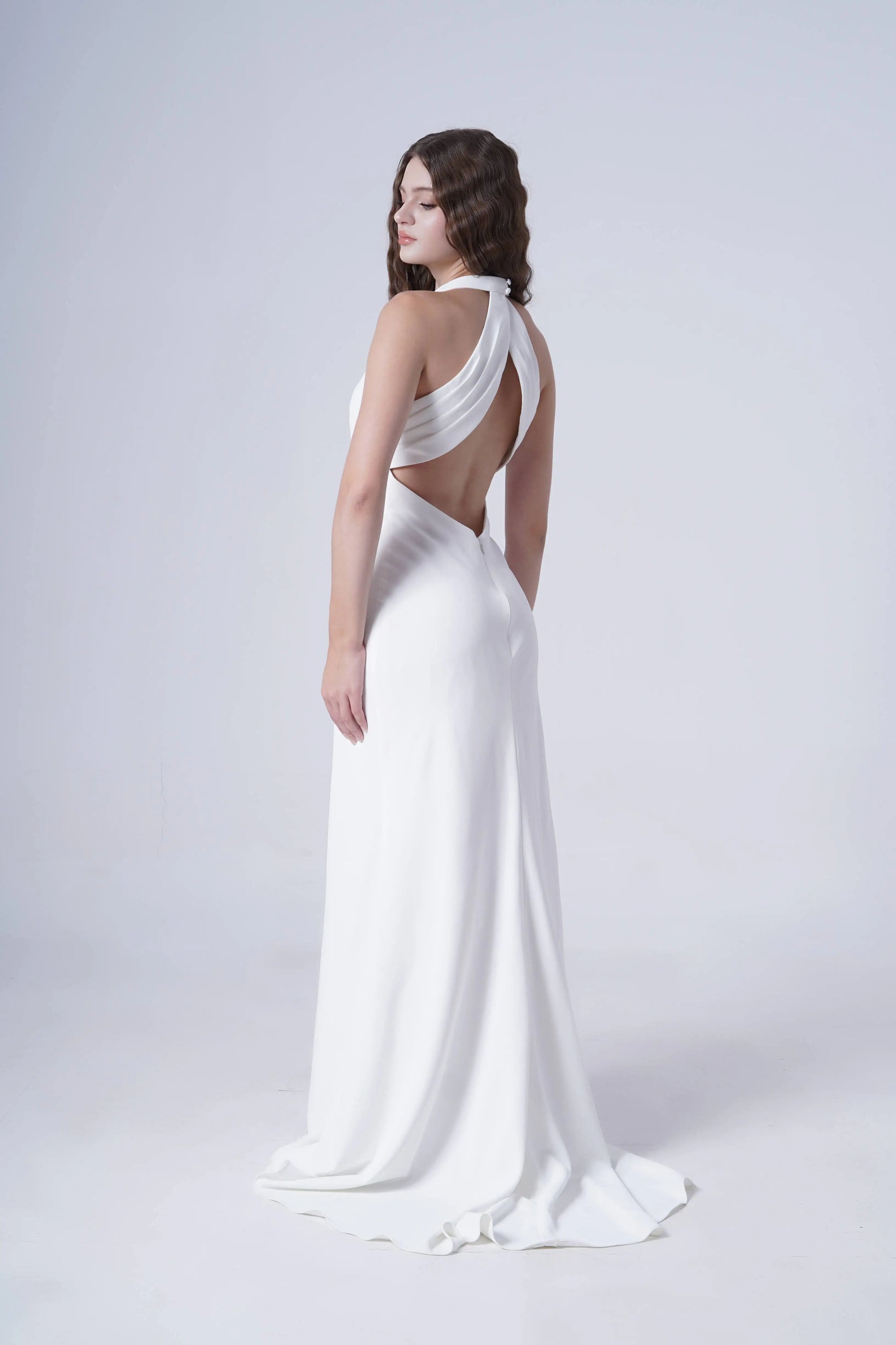 A woman models the Colette Bridal Dress in Ivory by TH&TH—a matte crepe, floor-length gown with a draped cross-strap halter neckline and open back. She poses against a plain light background, highlighting the dress’s elegant flowing silhouette.