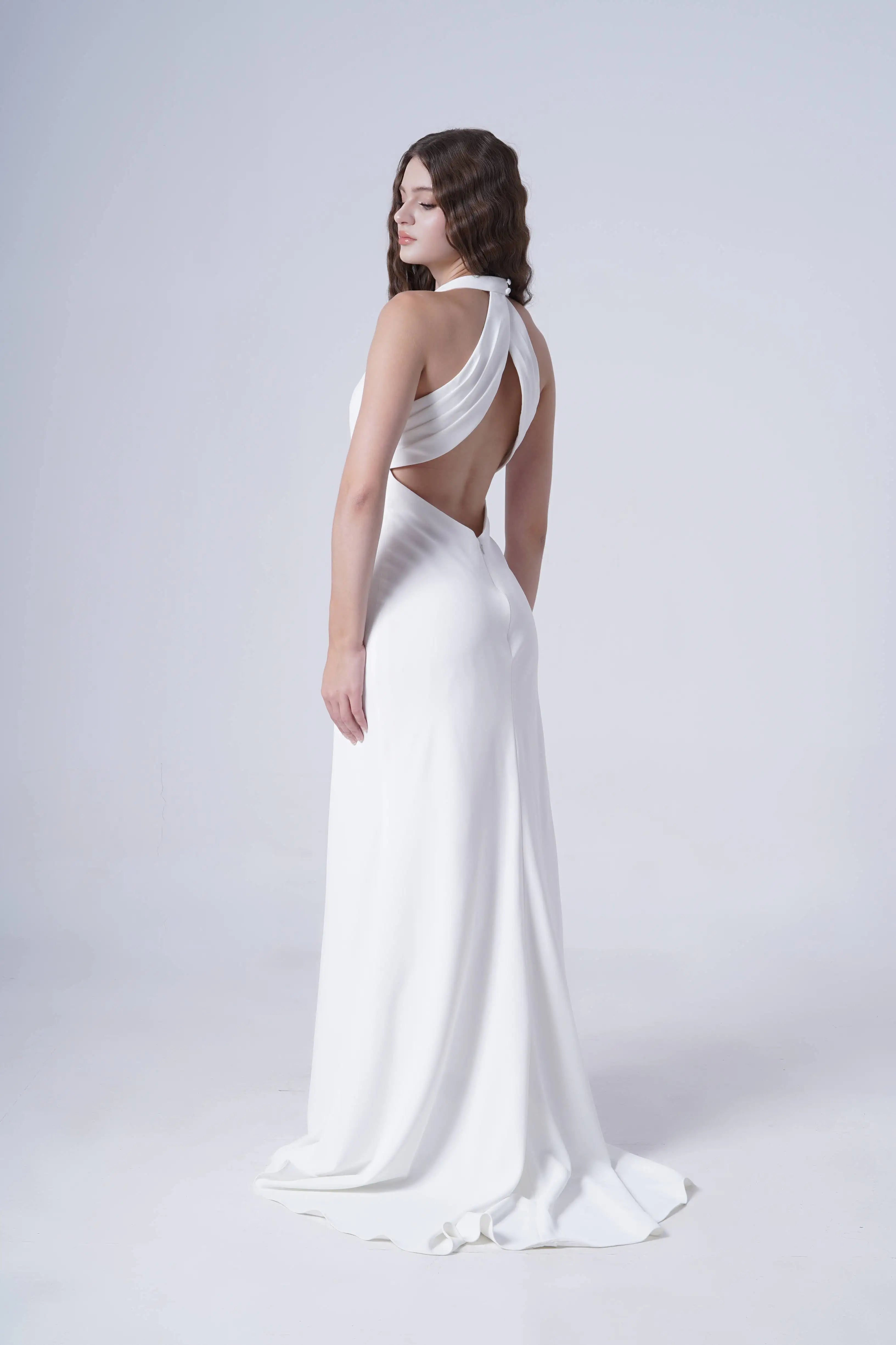 A woman models the Colette Bridal Dress in Ivory by TH&TH—a matte crepe, floor-length gown with a draped cross-strap halter neckline and open back. She poses against a plain light background, highlighting the dress’s elegant flowing silhouette.