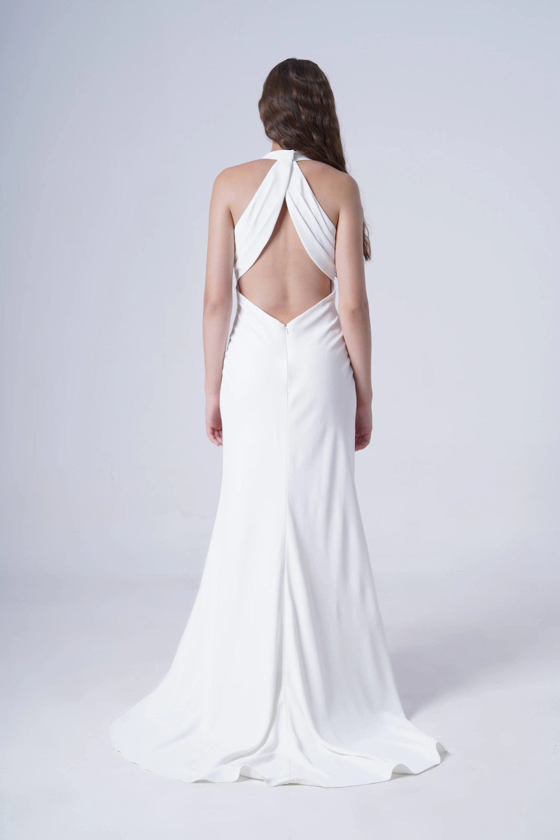 A woman stands facing away against a plain light background, wearing the Colette Bridal Dress in Ivory by TH&TH—a long bridal gown with a halter neckline, open back, and crisscross straps.