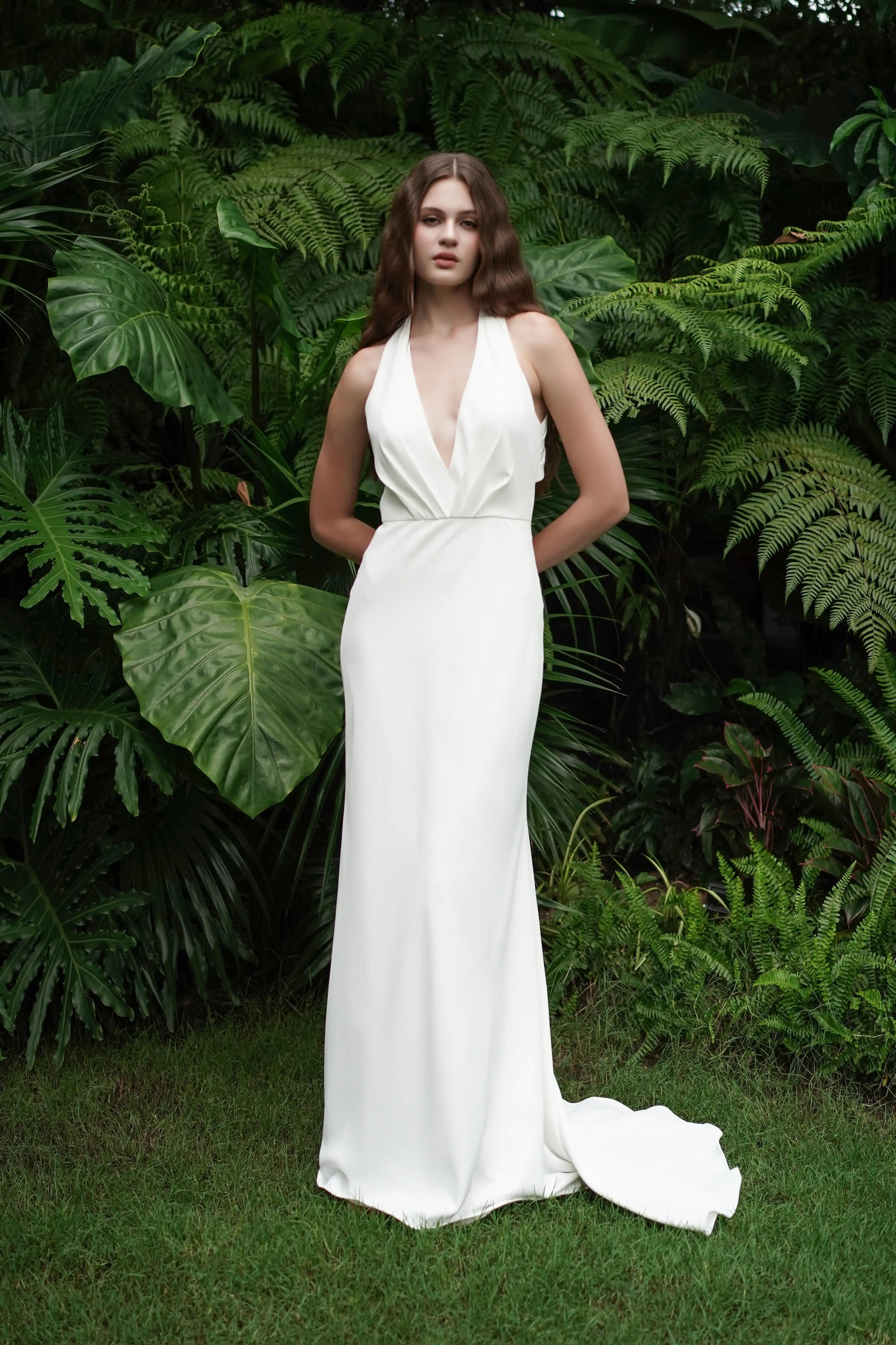 A woman wearing the TH&TH Colette Bridal Dress in Ivory, a sleeveless matt crepe gown with a halter neckline, stands on grass before lush tropical plants, facing the camera with her hands at her sides.