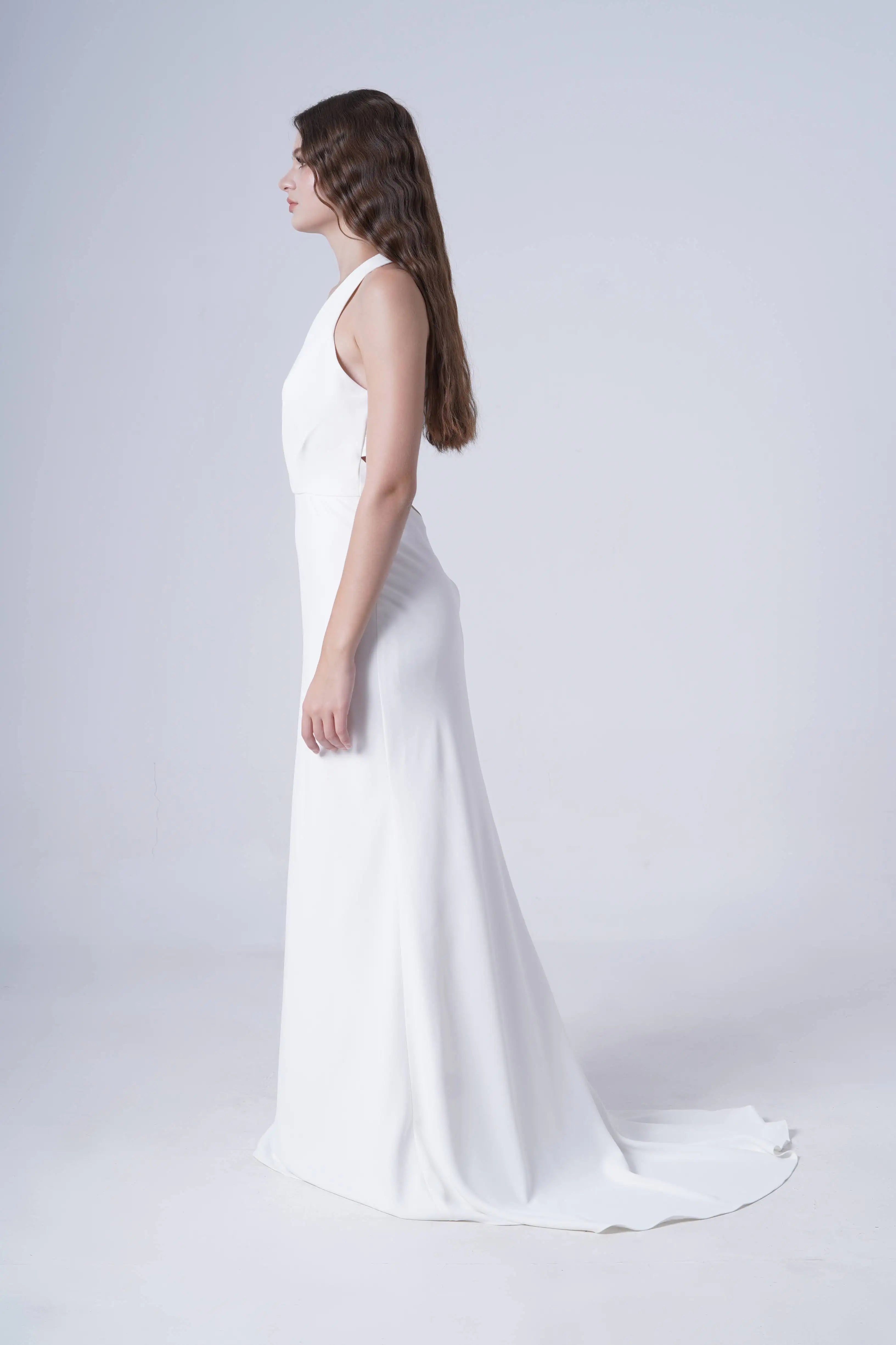 A woman with long brown hair stands in profile against a plain light gray background, wearing the sleeveless, floor-length Colette Bridal Dress in Ivory by TH&TH with a halter neckline.