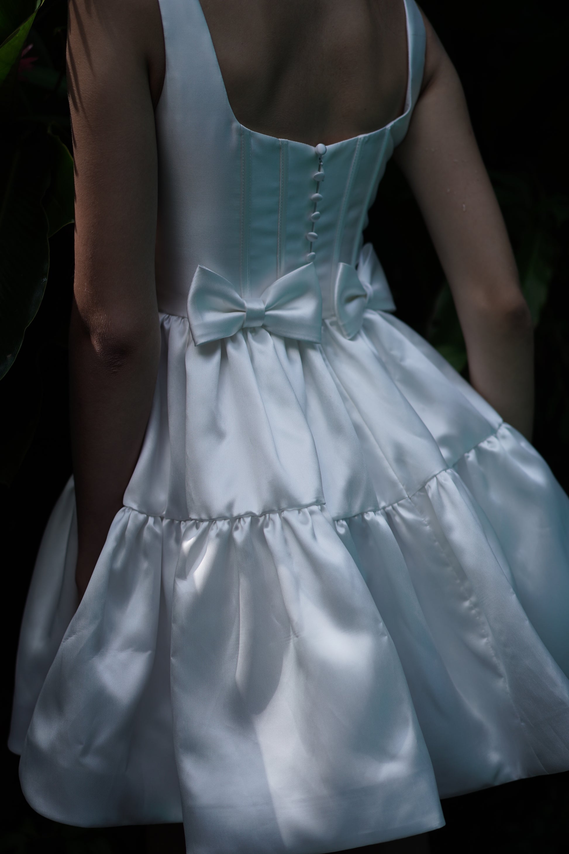 Wearing the TH&TH Bloom Bridal Mini Dress in Ivory, featuring a sleeveless satin twill design with a corseted bodice, buttoned back, bow at the waist, and full ruffled skirt, she stands in soft natural light.