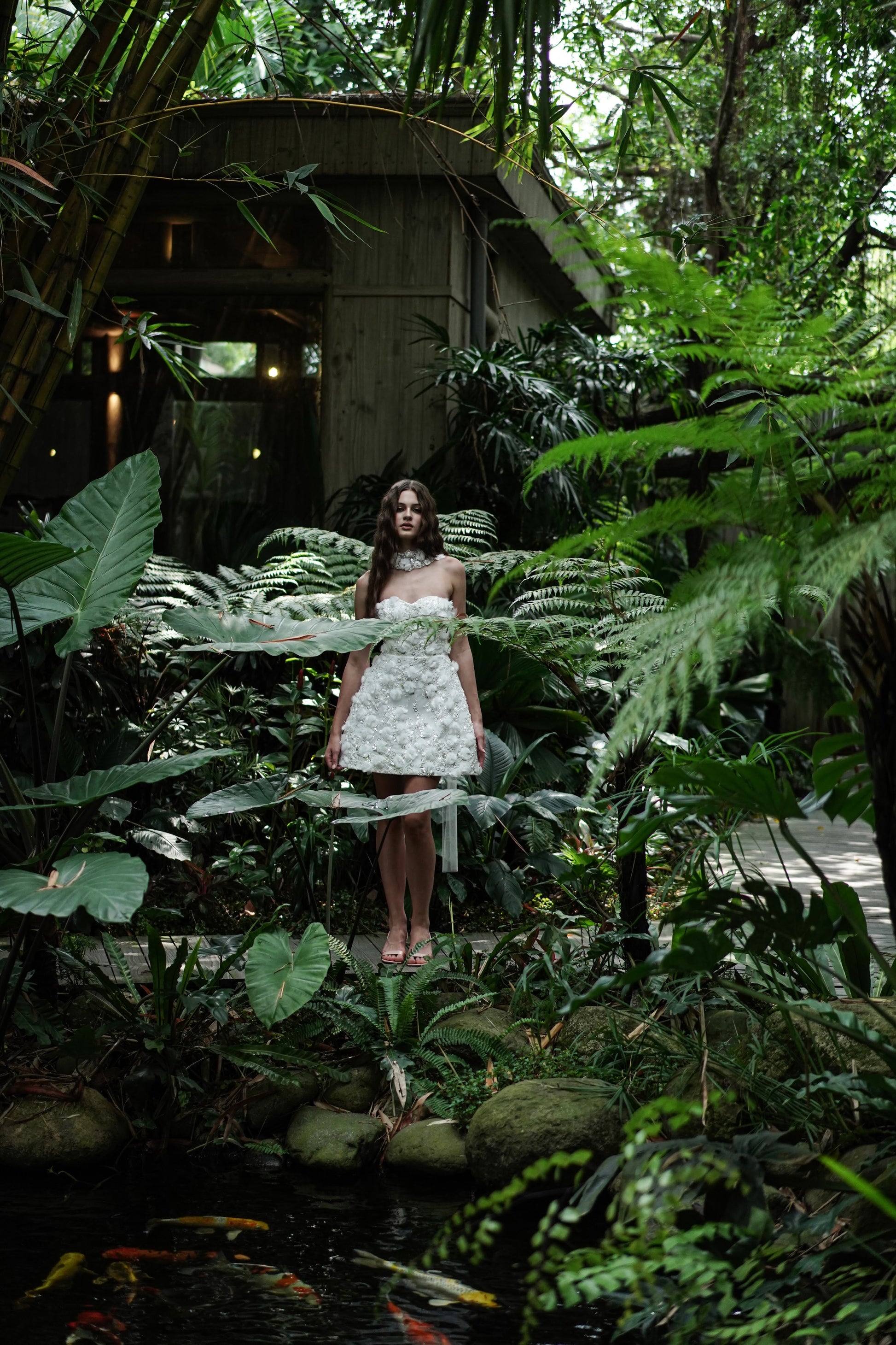 Woman in a white dress standing in a lush green forest with a building in the background.