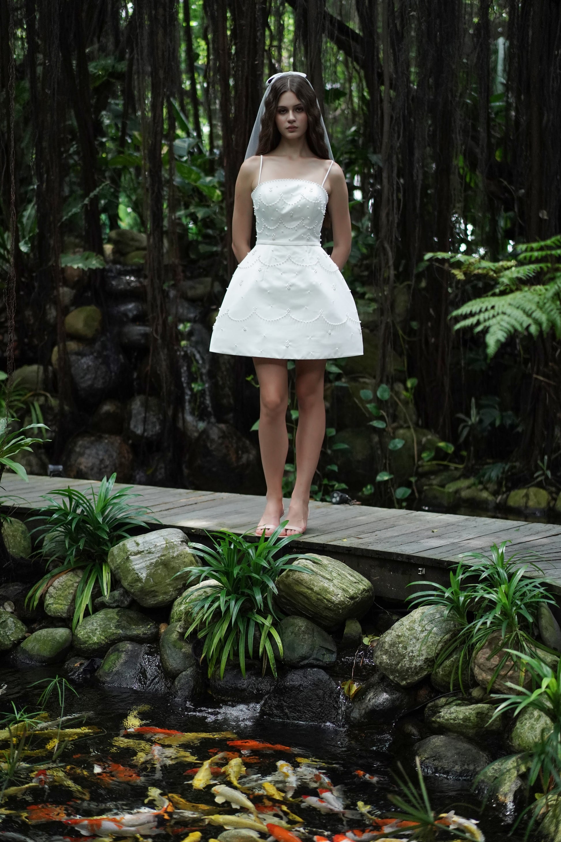 A woman wears the TH&TH Pearl Bridal Mini Dress in Ivory, standing barefoot on a wooden bridge in a lush garden beside rocks, greenery, and a pond with orange koi fish.