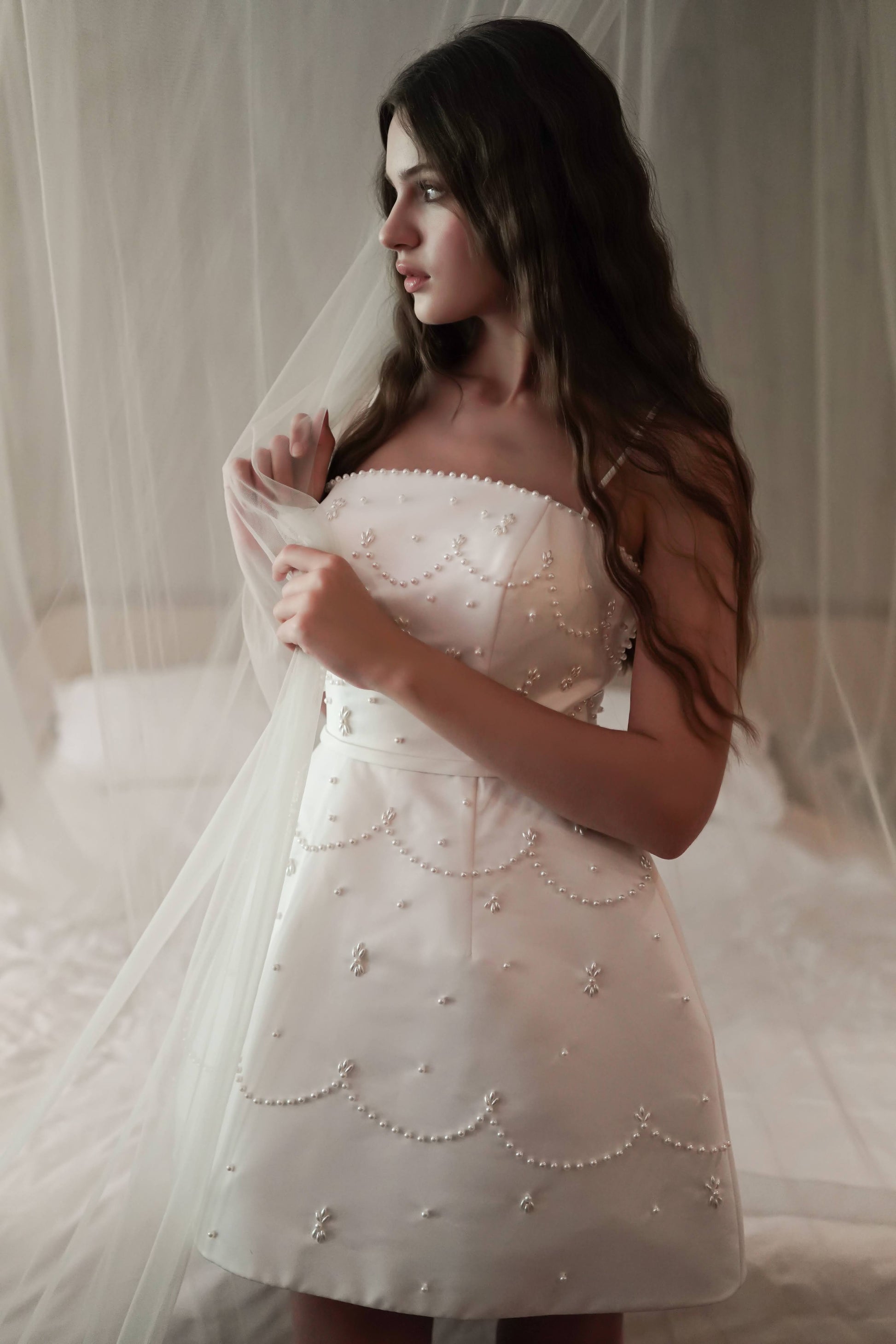 A young woman with long wavy hair stands indoors, wearing the TH&TH Pearl Bridal Mini Dress in Ivory and holding a sheer white veil, surrounded by soft lighting and delicate fabric drapery.