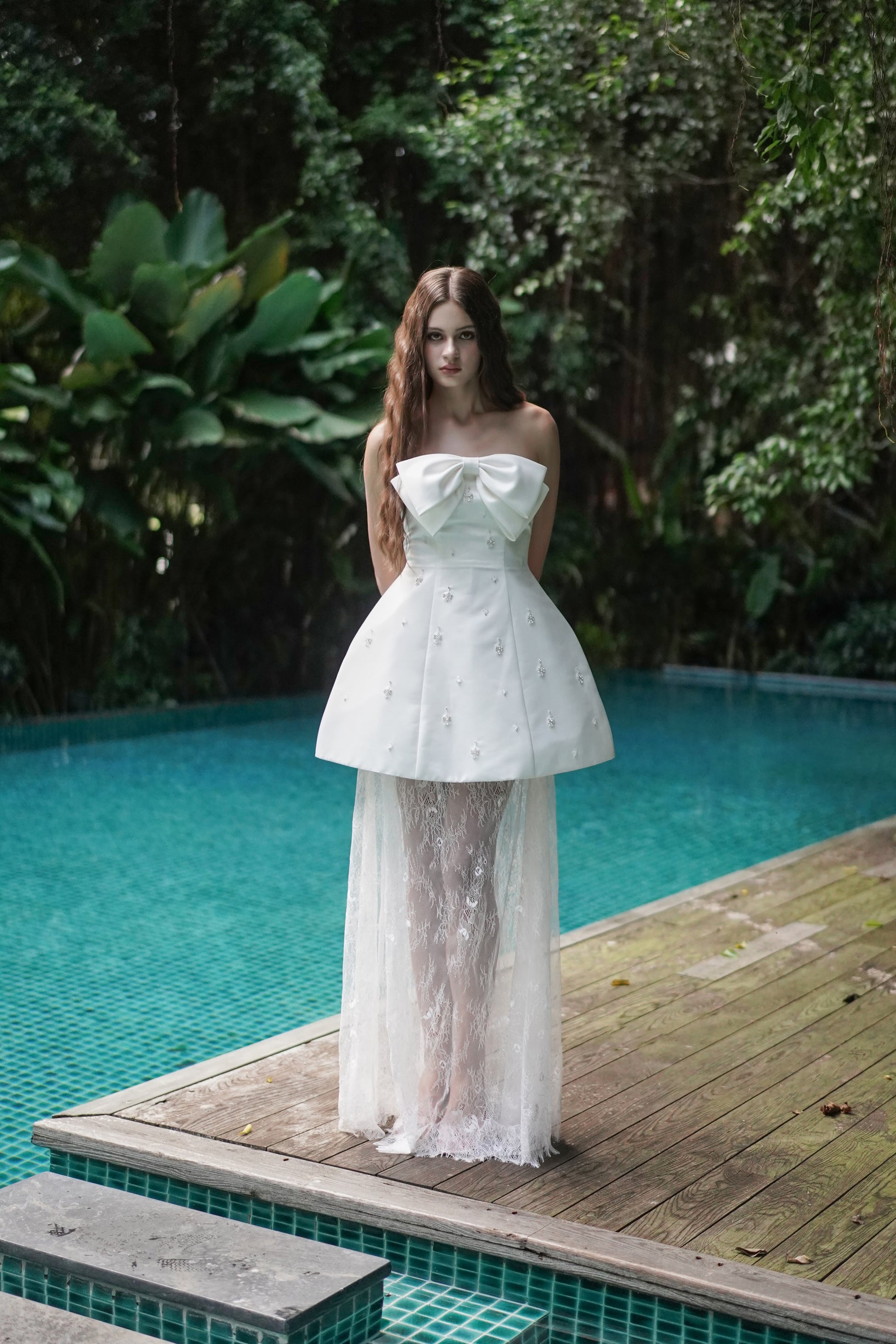 Woman in a white strapless dress standing by a pool with greenery in the background
