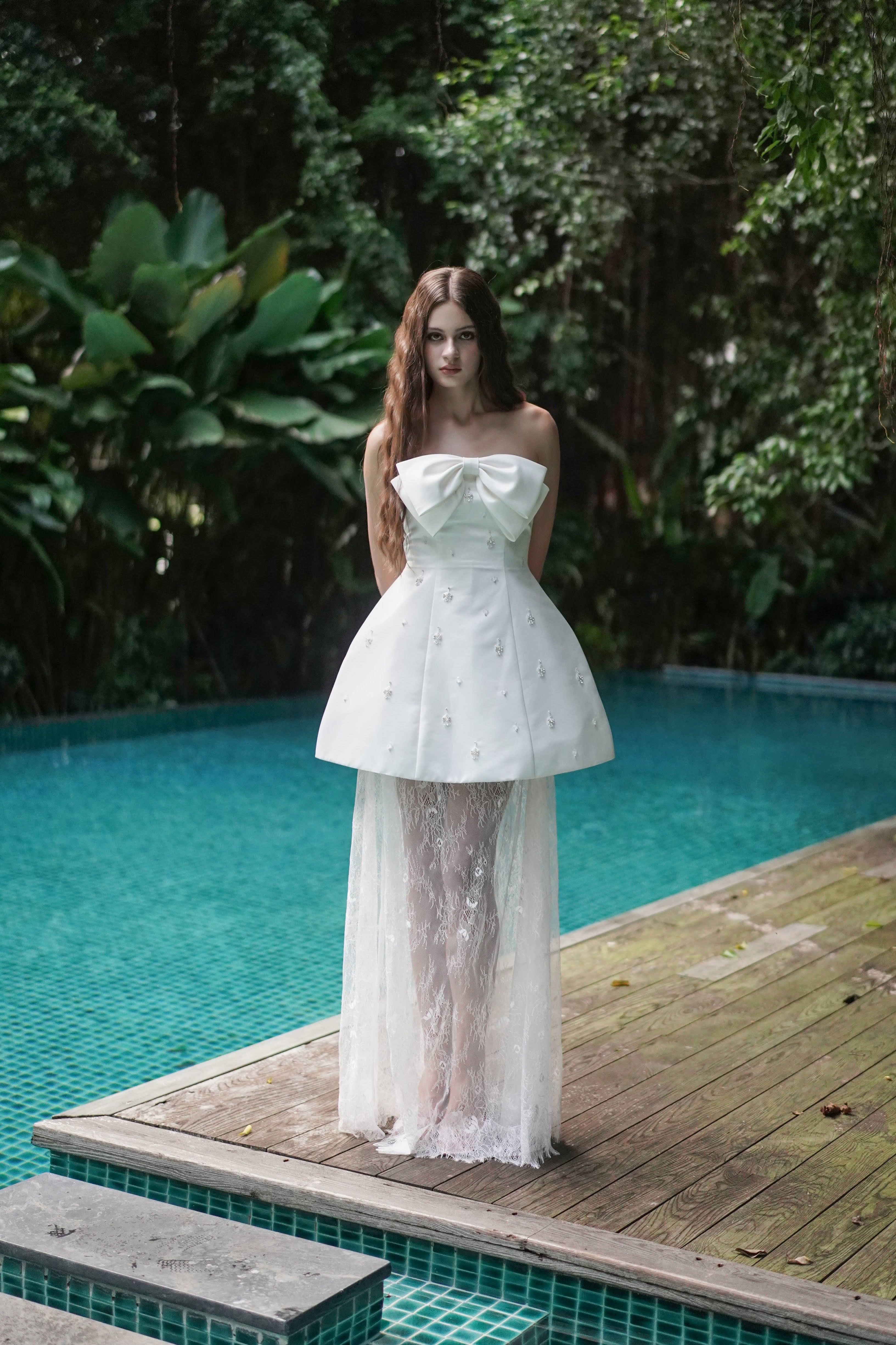 Woman in a white strapless dress standing by a pool with greenery in the background