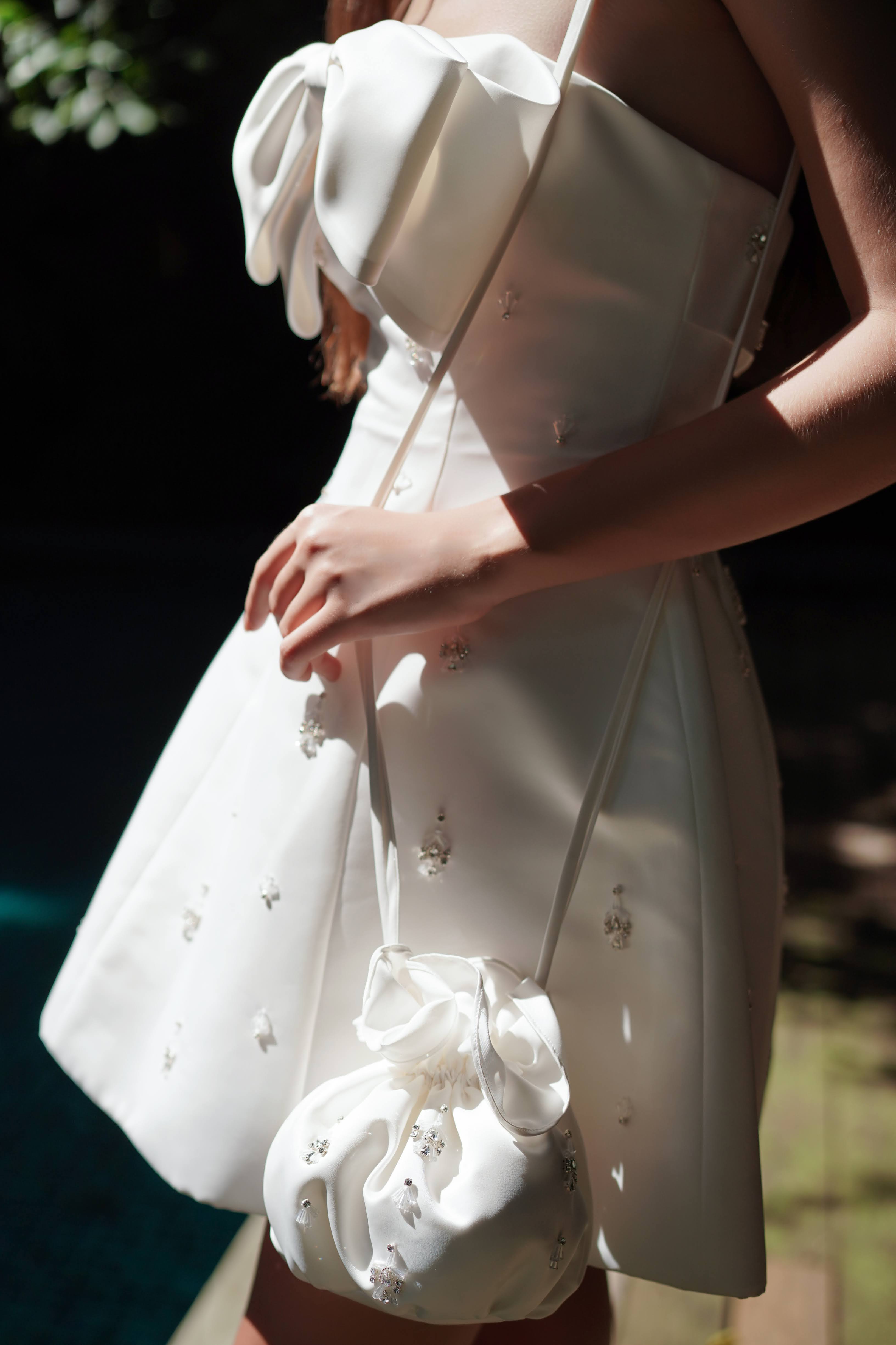 A woman in a sleeveless white dress with a large crystal-embellished bow holds the TH&TH 'Zephyr Crystal' Bridal Bag in Ivory, both sparkling with crystal details as sunlight enhances their shimmering beauty.