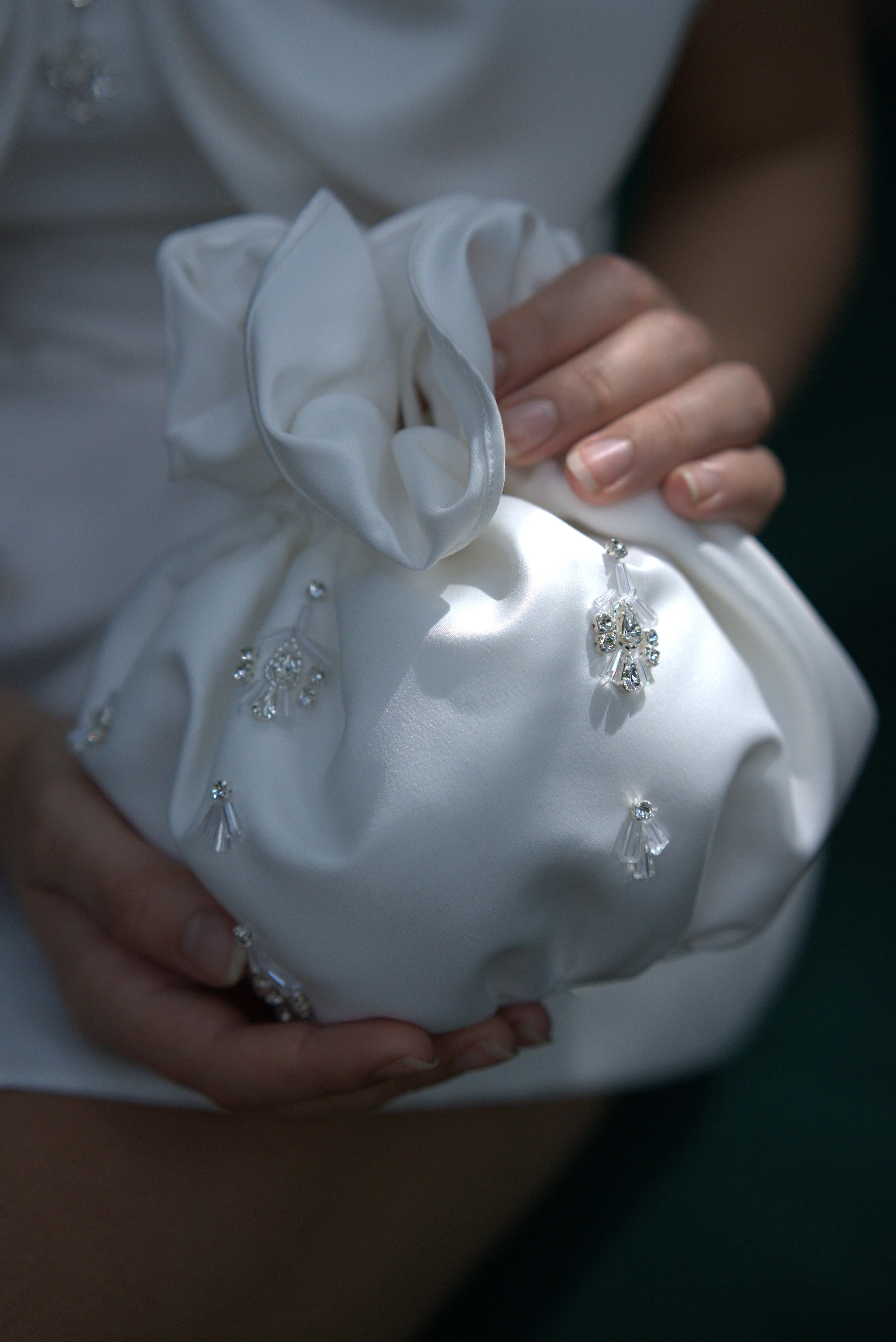 A person gently holds the TH&TH 'Zephyr Crystal' Bridal Bag in Ivory, its satin fabric gathered at the top and adorned with sparkling crystal embellishments for an elegant touch.
