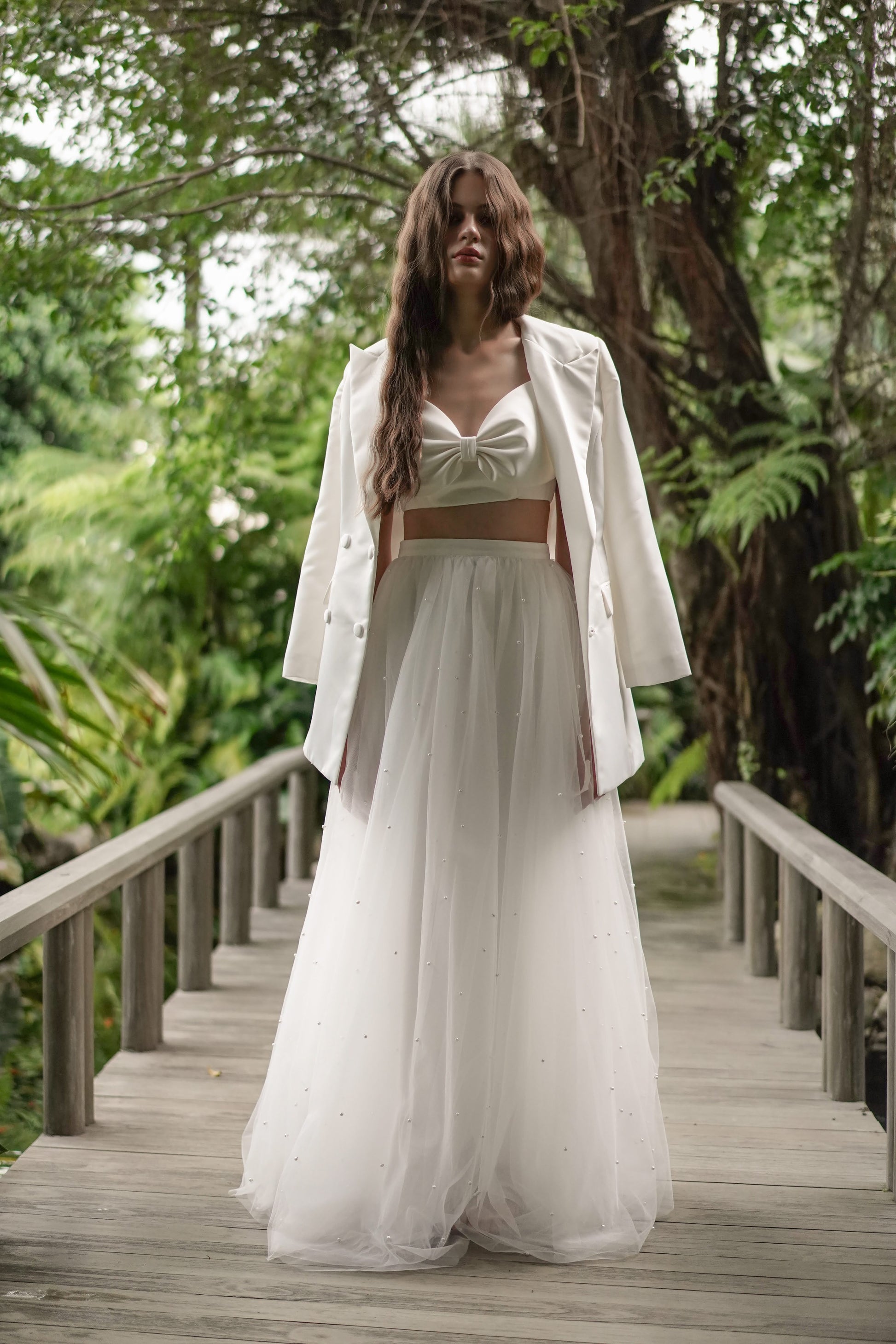 Woman in a white blazer standing on a wooden bridge with greenery in the background