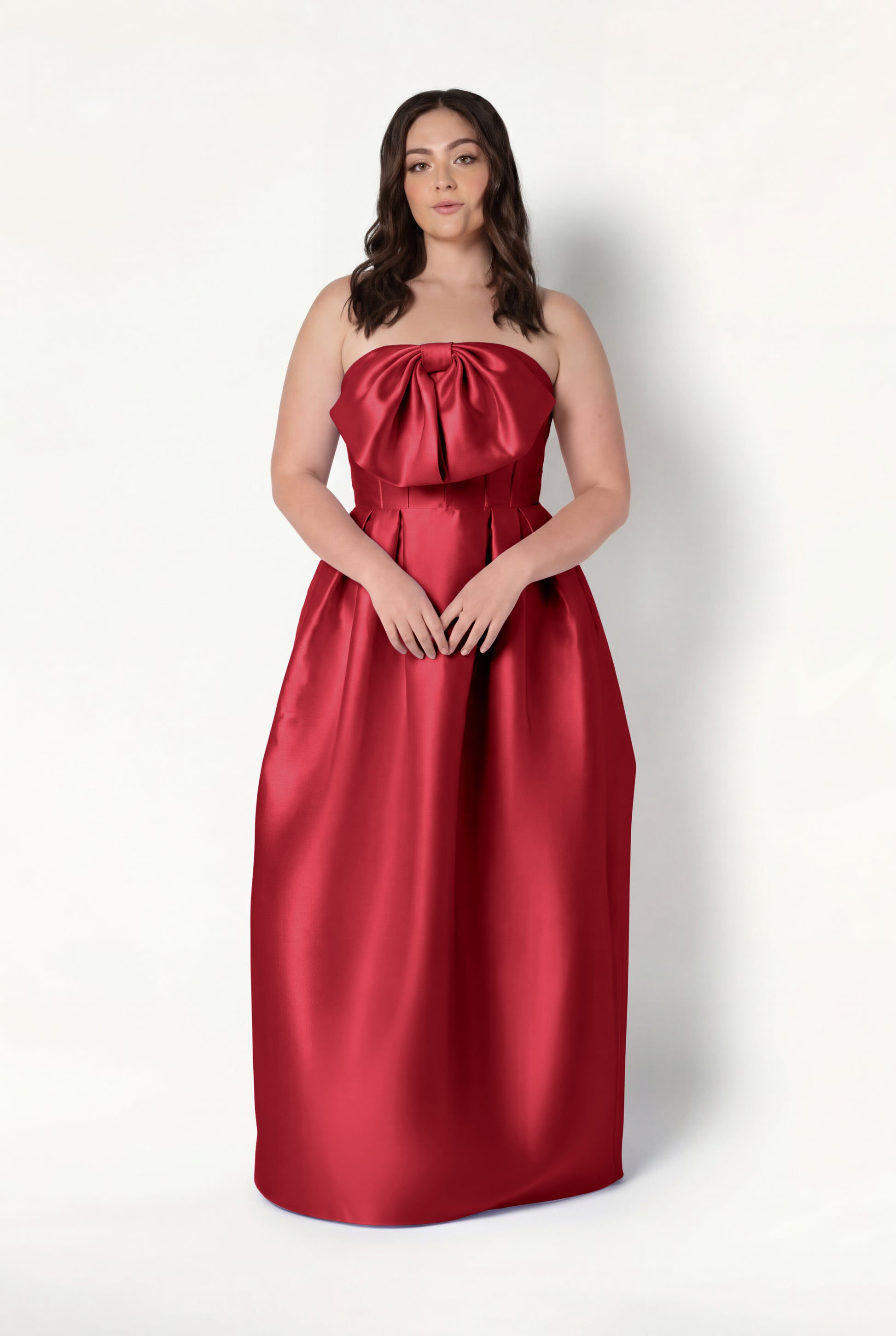 A woman with long brown hair models the Ophelia Bridesmaid Dress in Dark Cherry Satin Twill by TH&TH, a strapless, floor-length gown with a large corset bodice bow, standing against a plain white background.