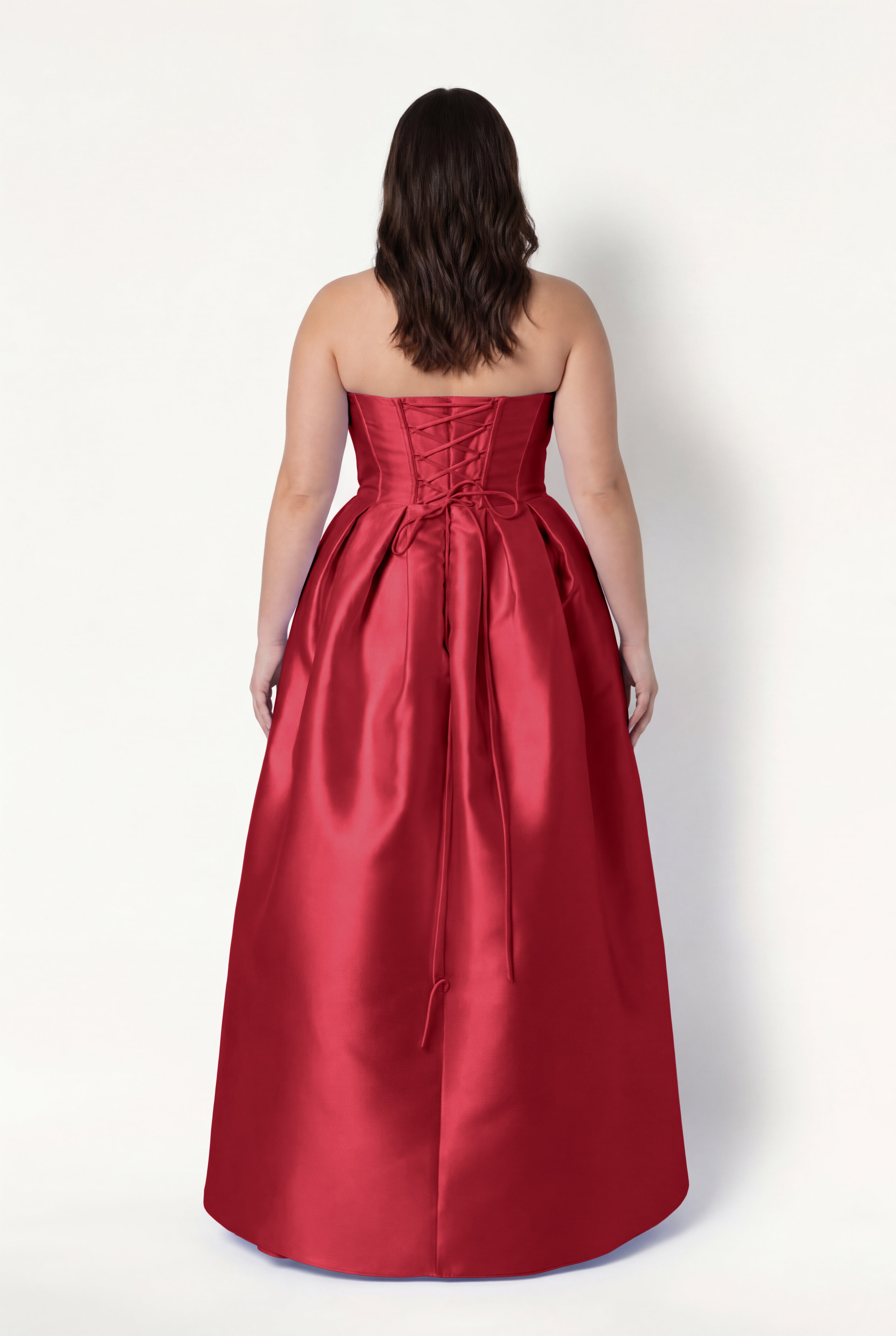 A woman with wavy brown hair faces away, wearing the TH&TH Ophelia Bridesmaid Dress in Dark Cherry Satin Twill—a strapless, floor-length gown with a lace-up corset bodice and bow detail, set against a plain white background.