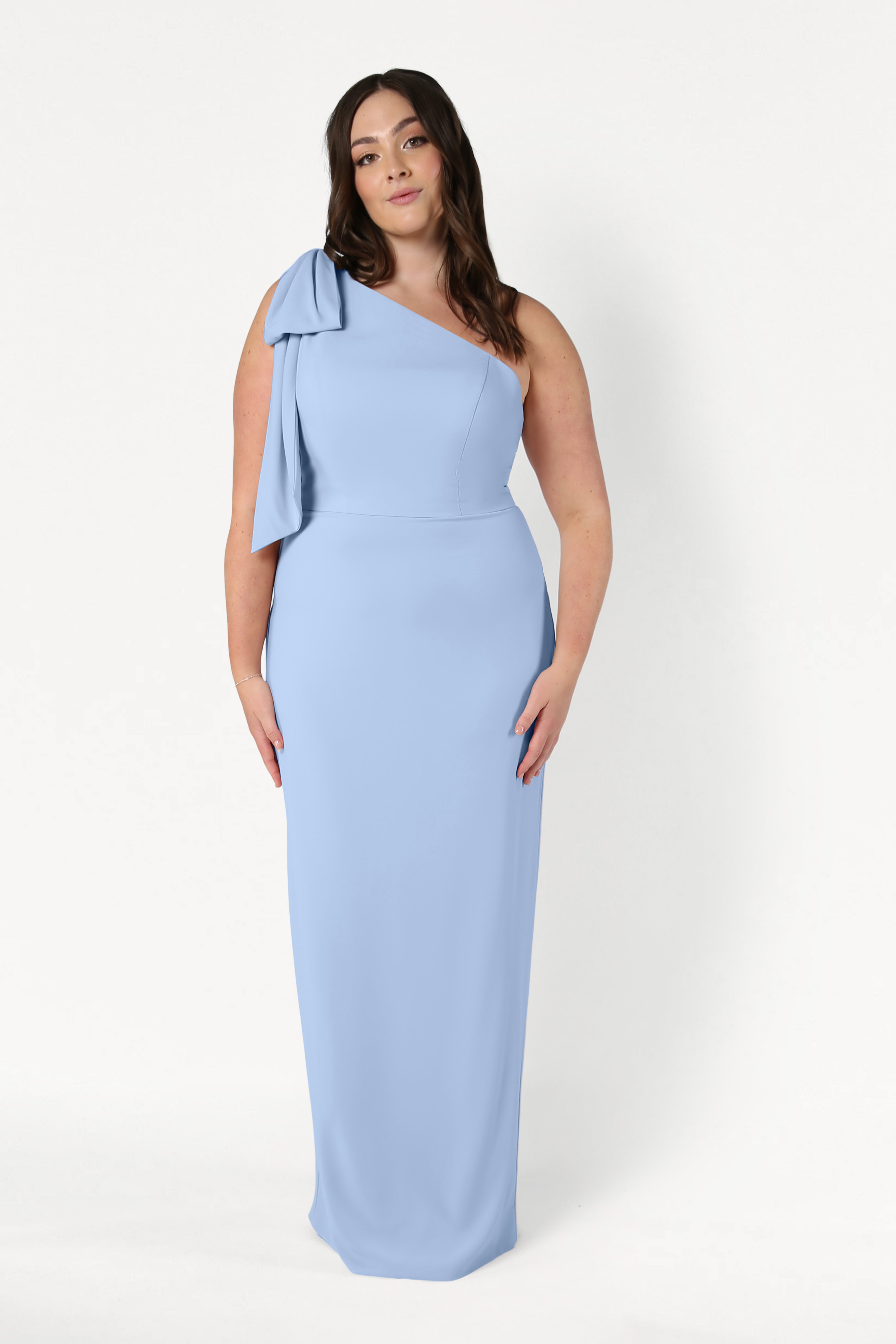 A woman stands against a white background wearing the TH&TH Valentina Bridesmaid Dress in Dusty Blue, a one-shoulder crepe gown featuring a bow detail on the shoulder.