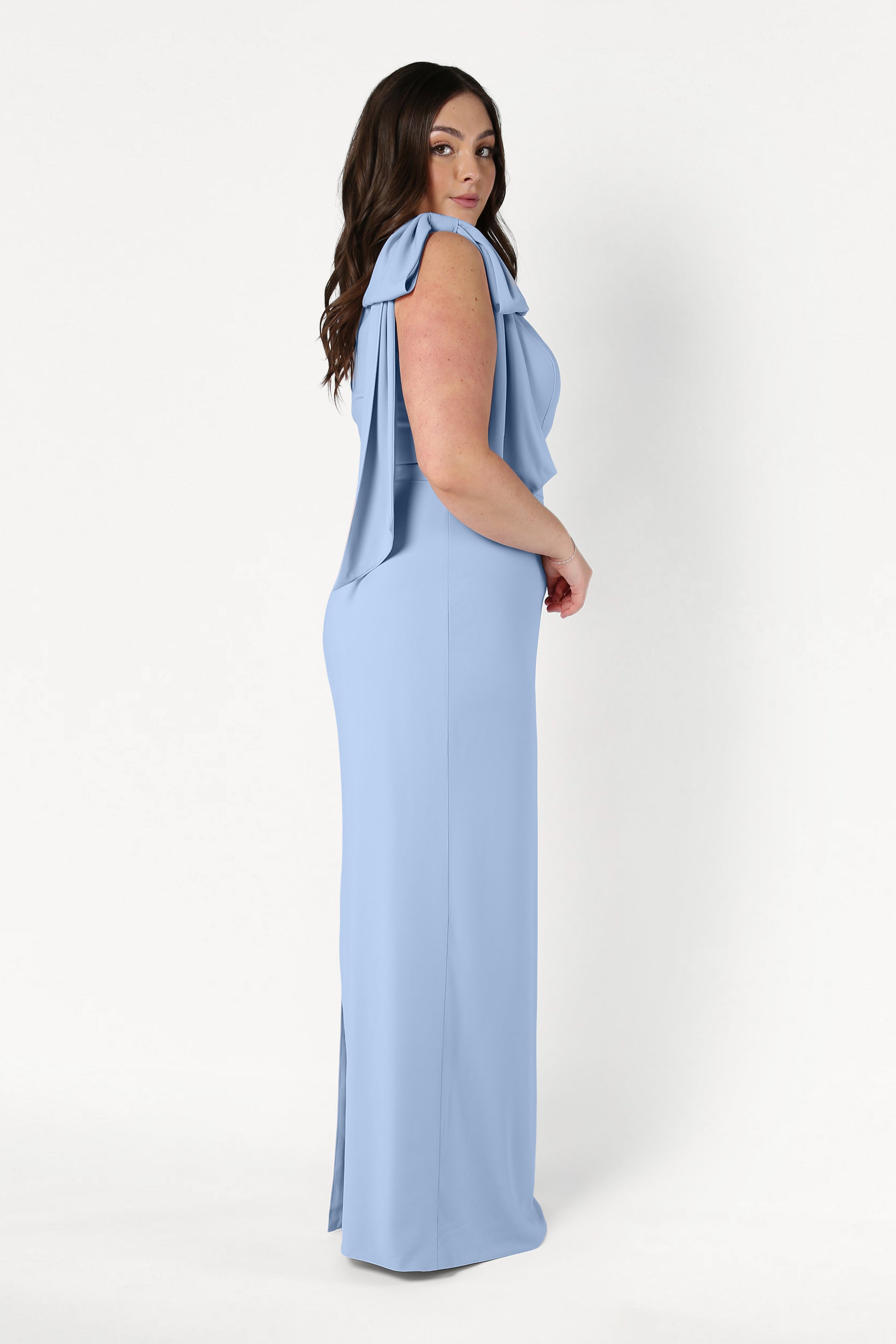 A woman with long, dark hair stands in profile wearing the TH&TH Valentina Bridesmaid Dress in Dusty Blue, featuring draped sleeves and a size-inclusive fit, set against a plain white background.