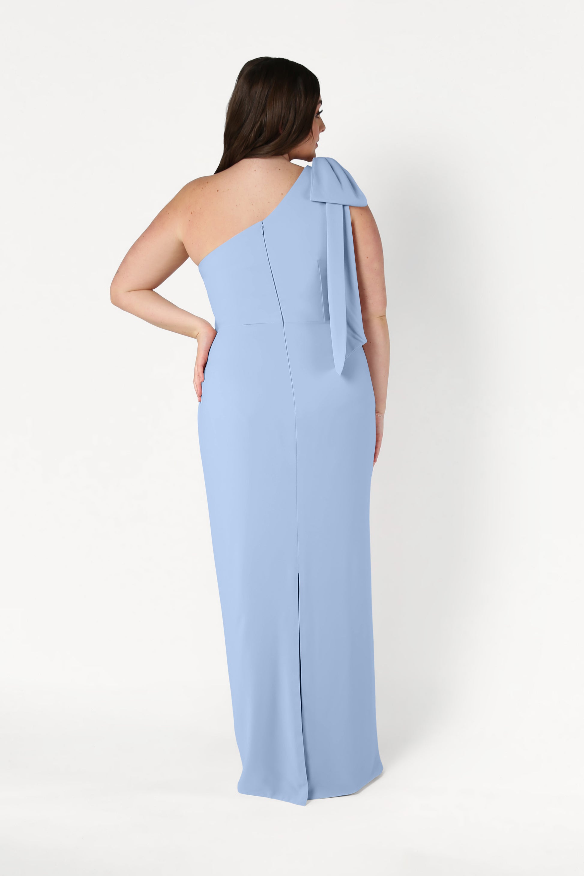 A woman faces away in the Valentina Bridesmaid Dress by TH&TH—a size-inclusive, floor-length, one-shoulder gown in dusty blue crepe with a bow detail, fitted silhouette, and subtle back slit—set against a plain white background.