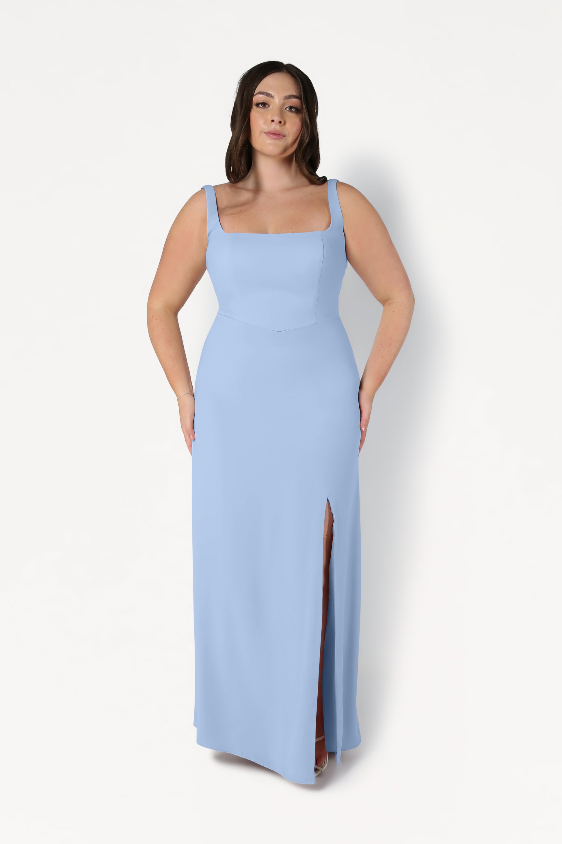 A woman stands against a white background wearing the Helena Bridesmaid Dress in Dusty Blue by TH&TH—a size-inclusive gown with a square neckline and front slit. She looks forward confidently, her hands at her sides.