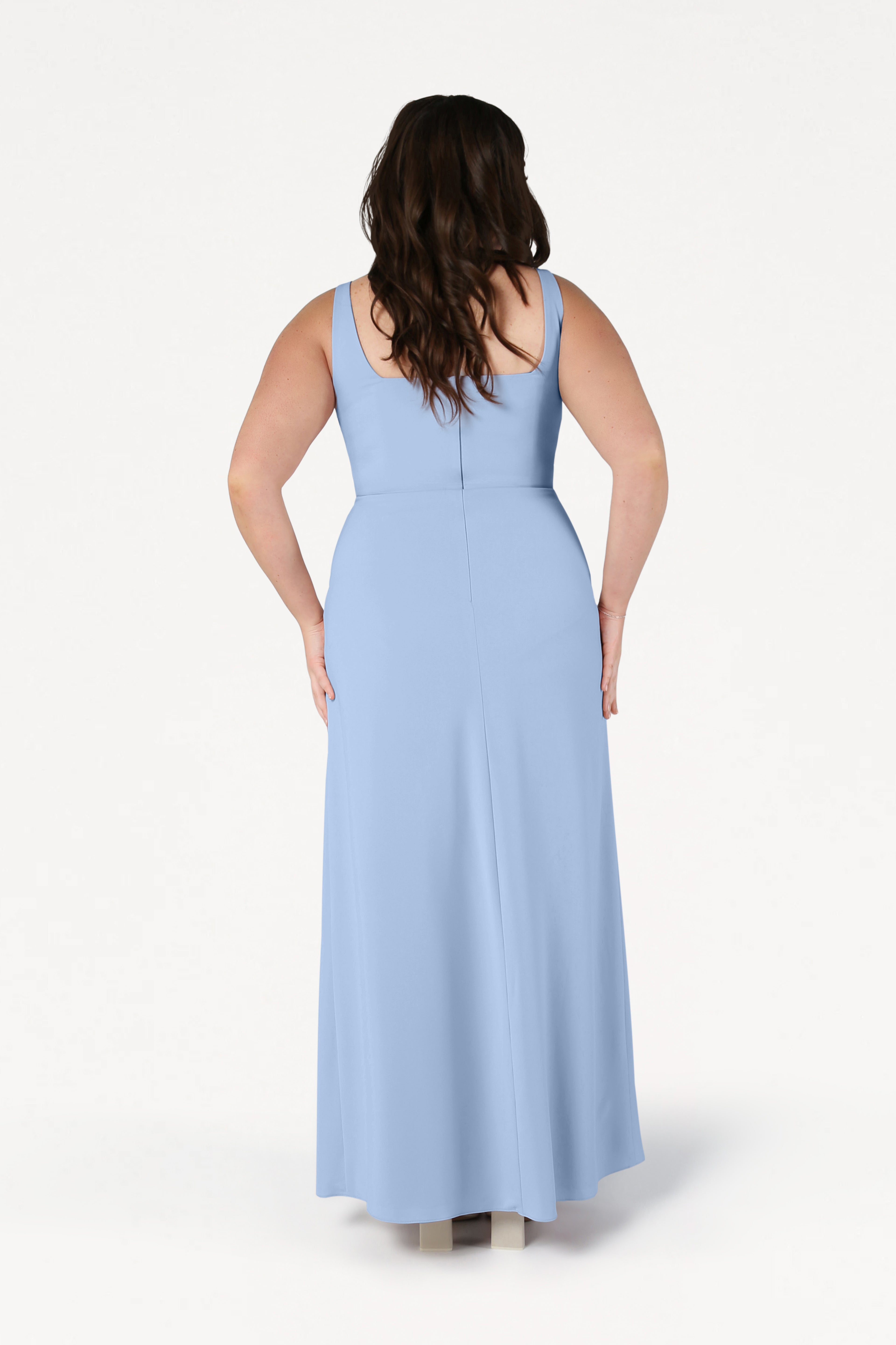 A woman with long brown hair stands facing away, wearing the TH&TH Helena Bridesmaid Dress in Dusty Blue and beige shoes against a plain white background.