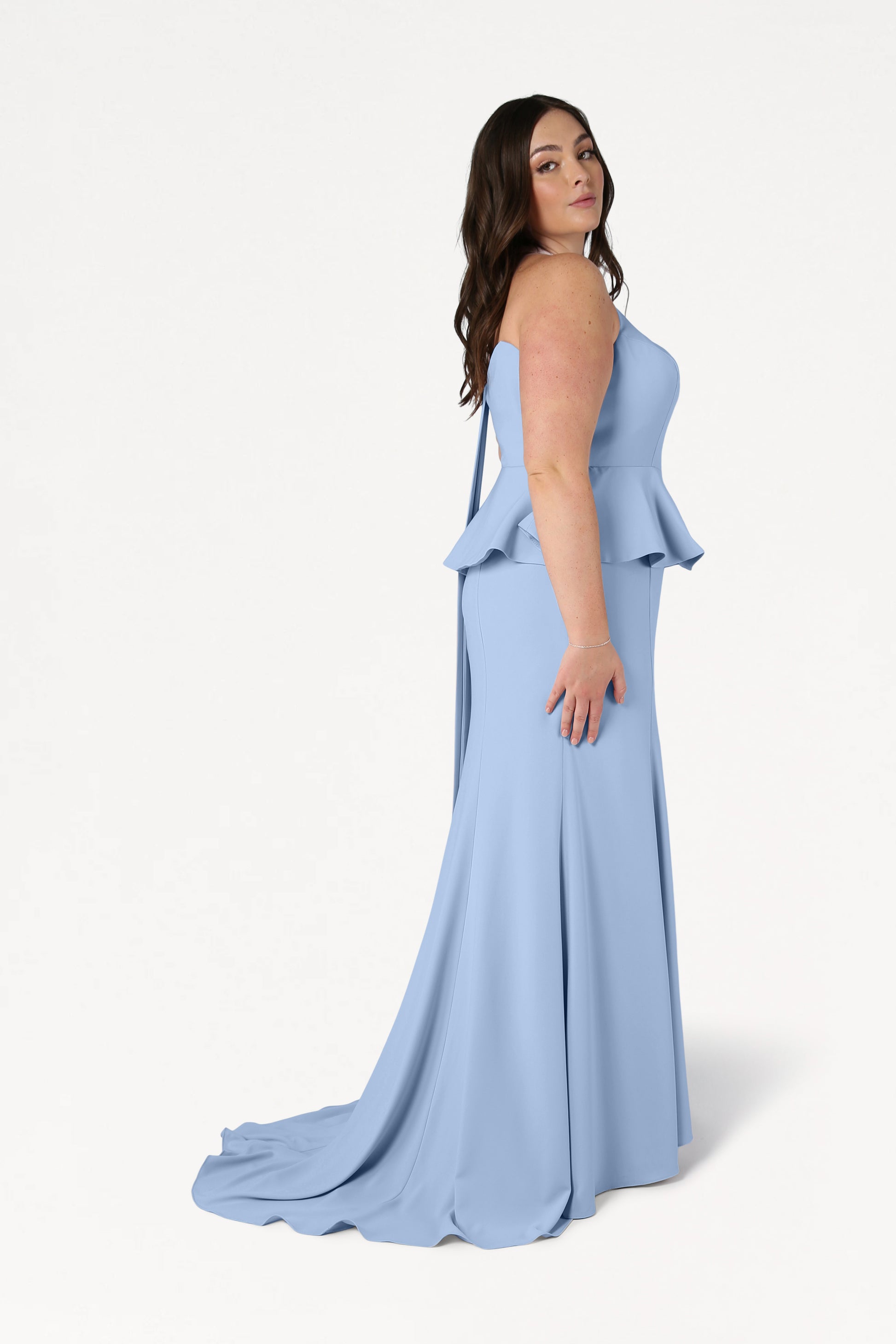 A woman with long brown hair wears the Peony Bridesmaid Dress in Dusty Blue by TH&TH, a modern, strapless, floor-length gown with peplum detail. This size-inclusive dress stands out as she confidently poses against a plain white background.