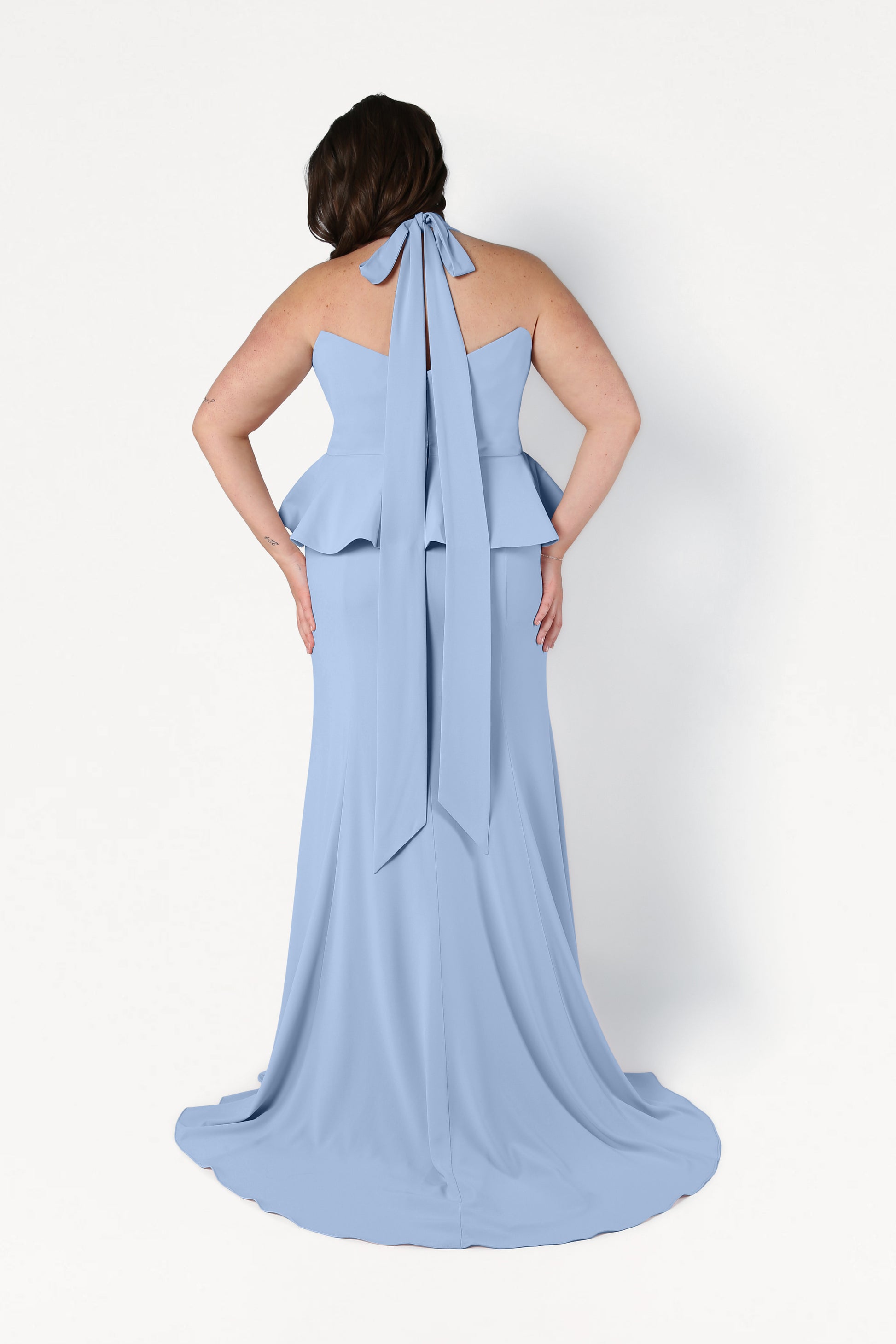A woman stands facing away in the TH&TH Peony Bridesmaid Dress in Dusty Blue, featuring a peplum waist, back bow tie, and flowing train—a chic, size-inclusive gown against a plain white background.