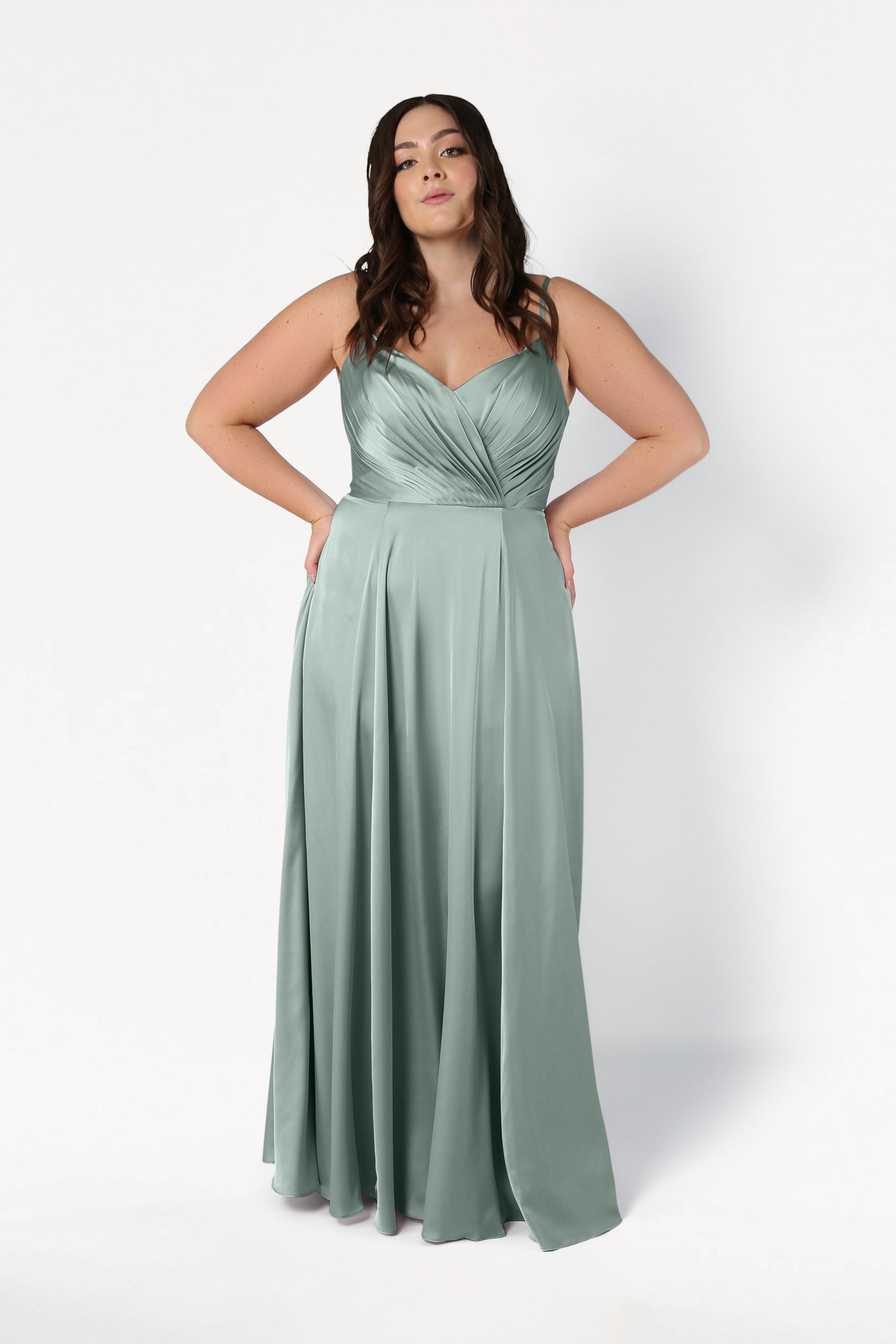 A woman with long brown hair wears the TH&TH Isla Bridesmaid Dress in Dusty Eucalyptus Satin, featuring a pleated bodice and flowing skirt. She stands against a plain white background with her hands on her hips.