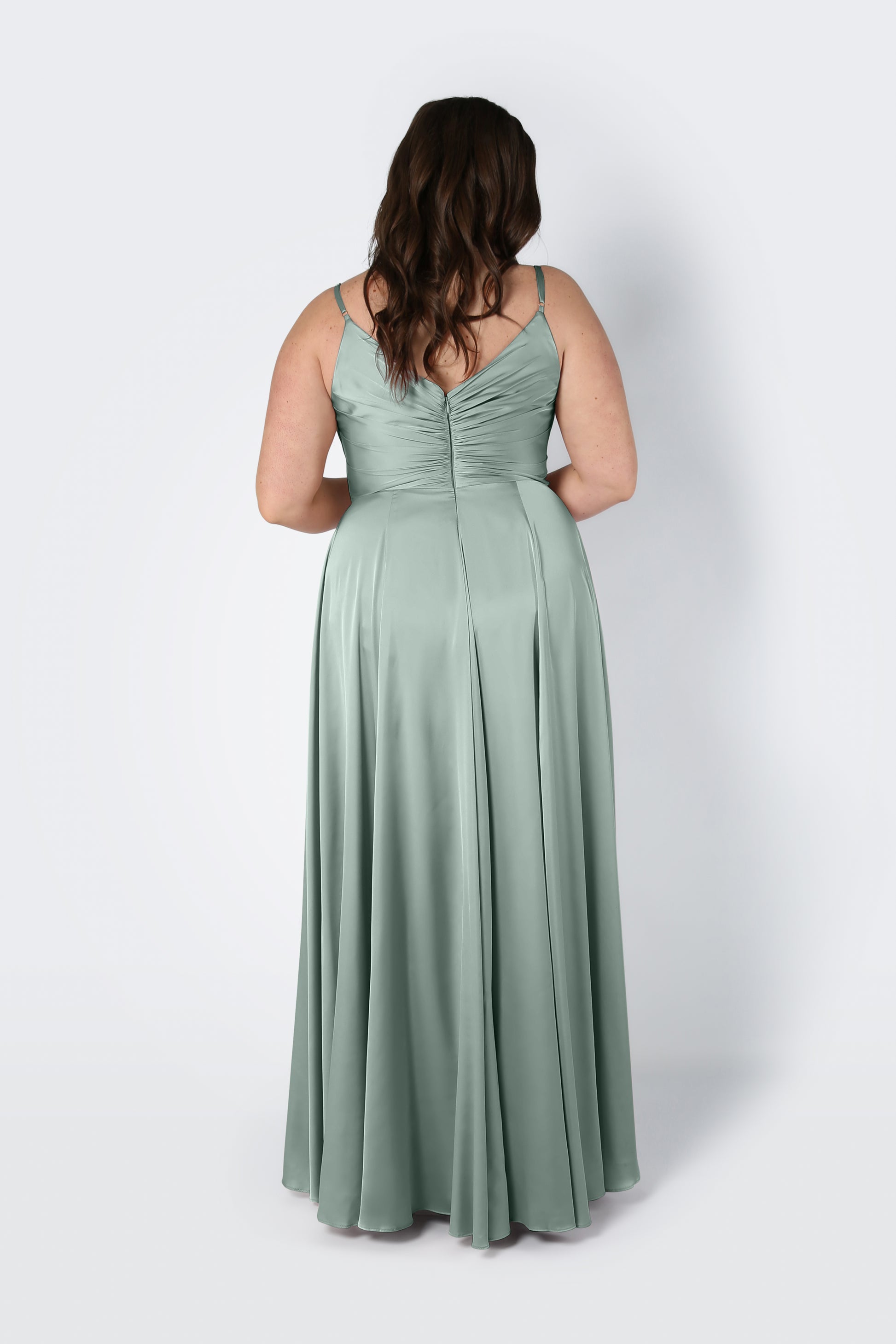 A woman with long brown hair wears the TH&TH Isla Bridesmaid Dress in Dusty Eucalyptus Satin, a floor-length gown with thin straps, pleated bodice, and ruched back, standing against a plain white background facing away from the camera.