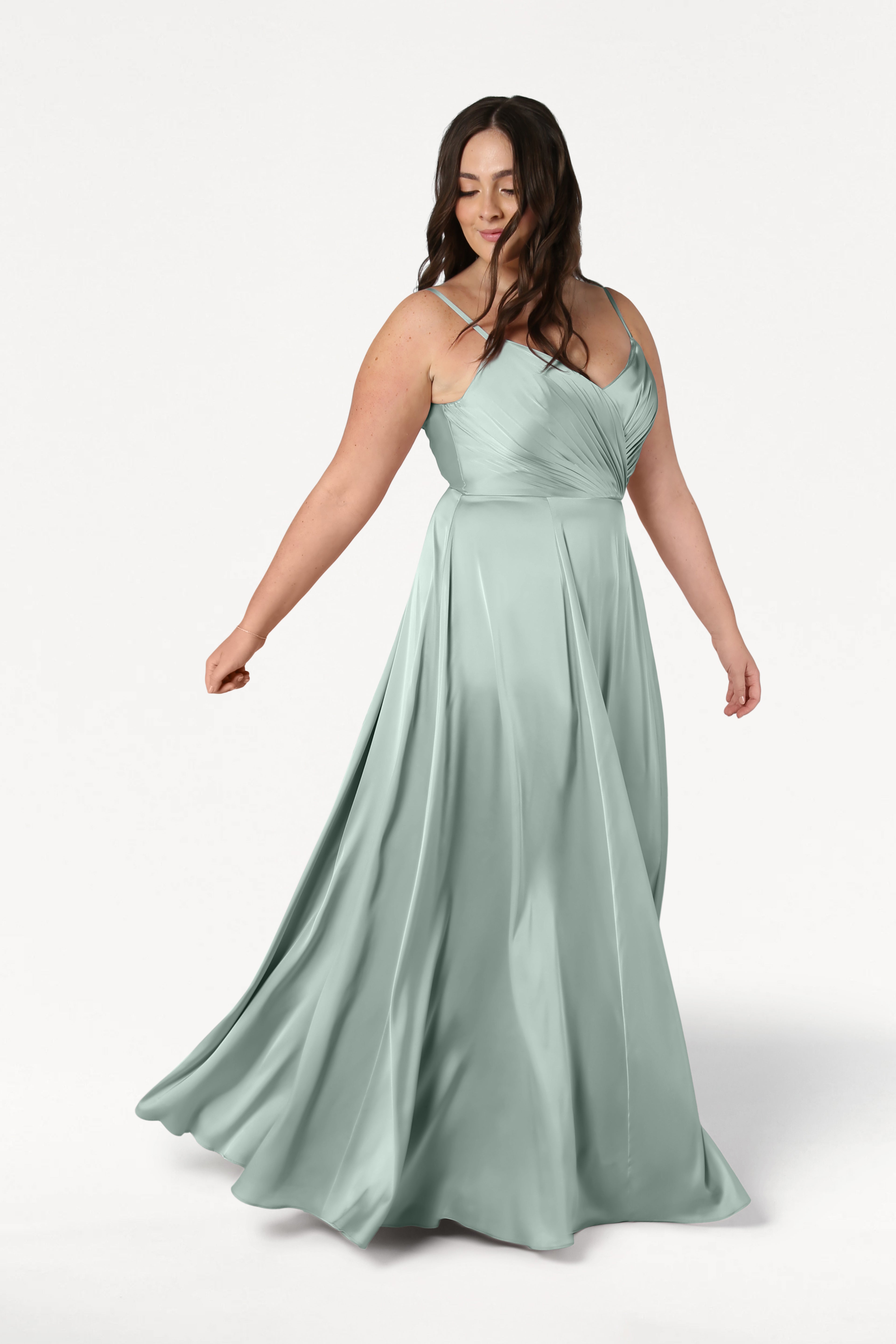 A woman with long brown hair twirls gracefully in a TH&TH Isla Bridesmaid Dress in Dusty Eucalyptus Satin, featuring a pleated bodice and floor-length skirt. The background is plain white.