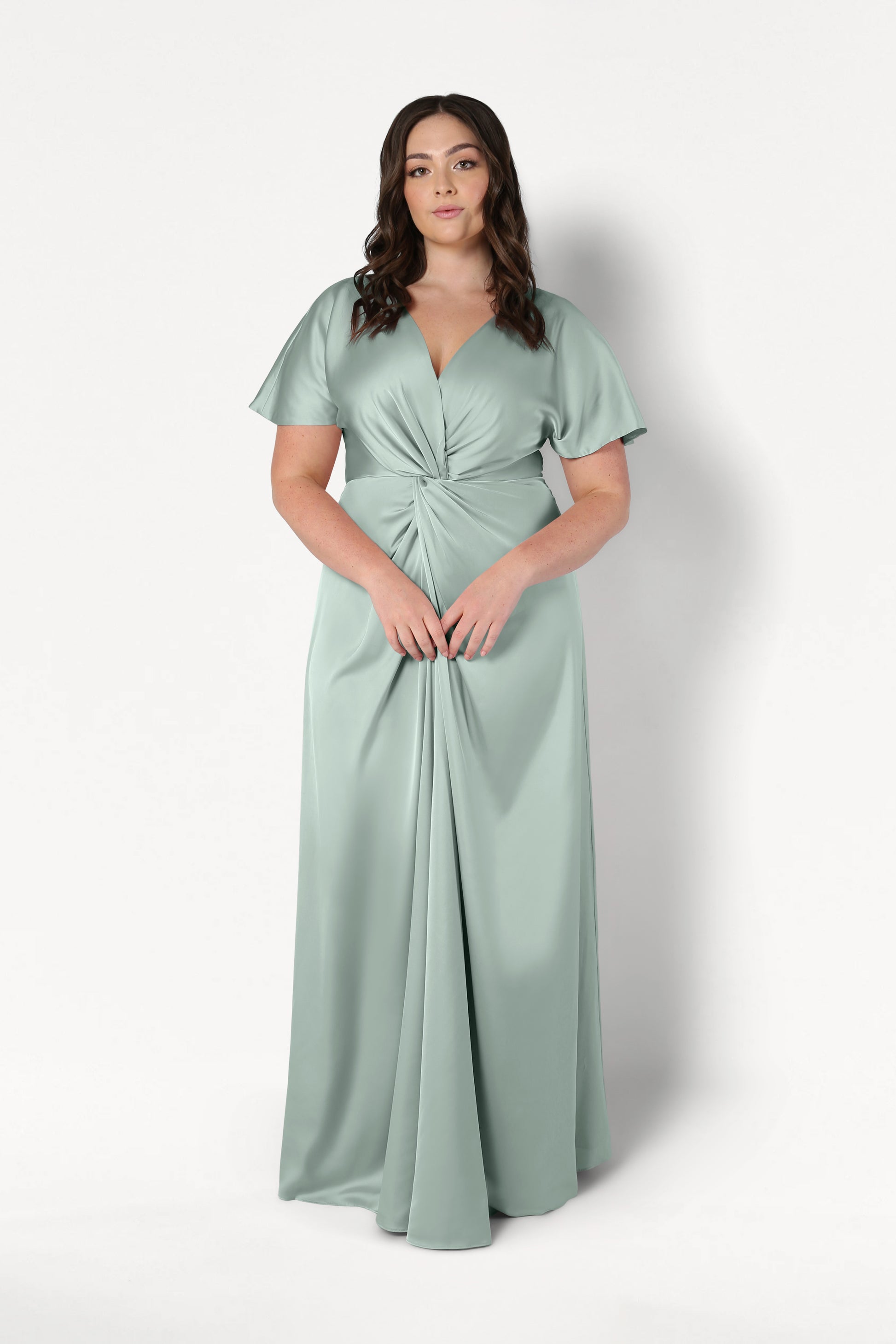 A woman poses against a white background wearing the TH&TH Camilla Bridesmaid Dress in Dusty Eucalyptus Satin, a floor-length gown with short flutter sleeves, a V-neckline, and a gathered twist-knot bodice at the waist.