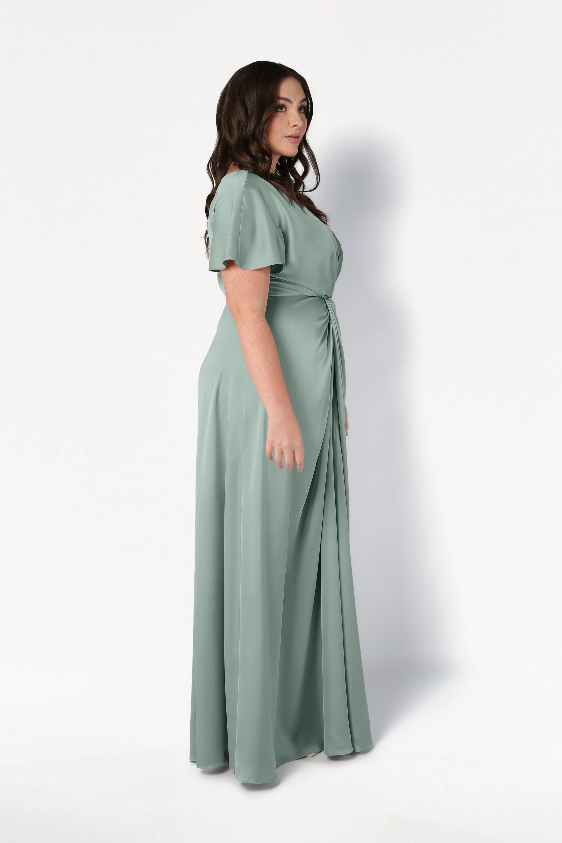 A woman with long brown hair stands in profile wearing the TH&TH Camilla Bridesmaid Dress in Dusty Eucalyptus Satin, a size-inclusive, floor-length design with short sleeves and a gathered waist, against a plain white background.