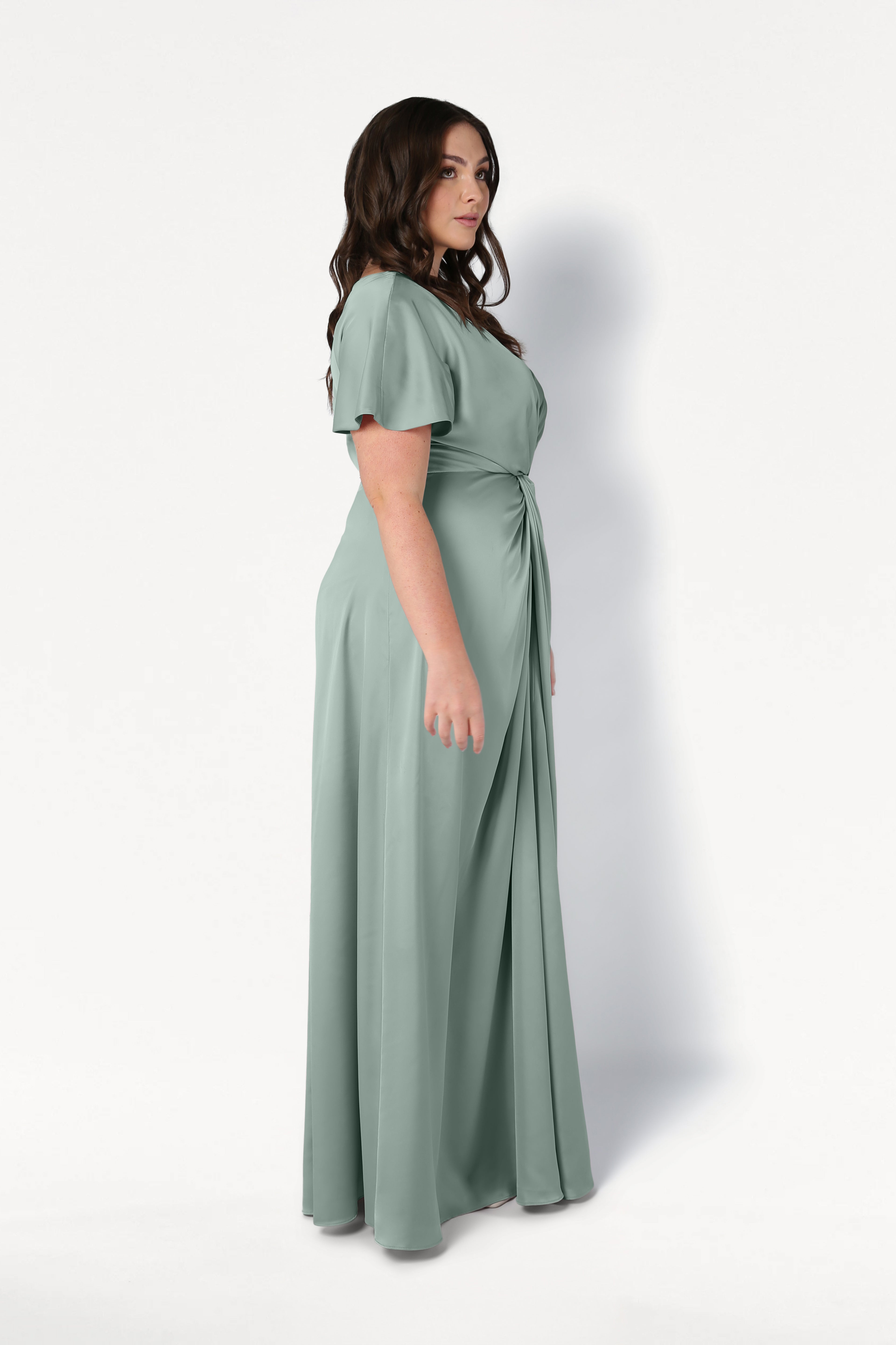 A woman with long brown hair stands in profile wearing the TH&TH Camilla Bridesmaid Dress in Dusty Eucalyptus Satin, a size-inclusive, floor-length design with short sleeves and a gathered waist, against a plain white background.