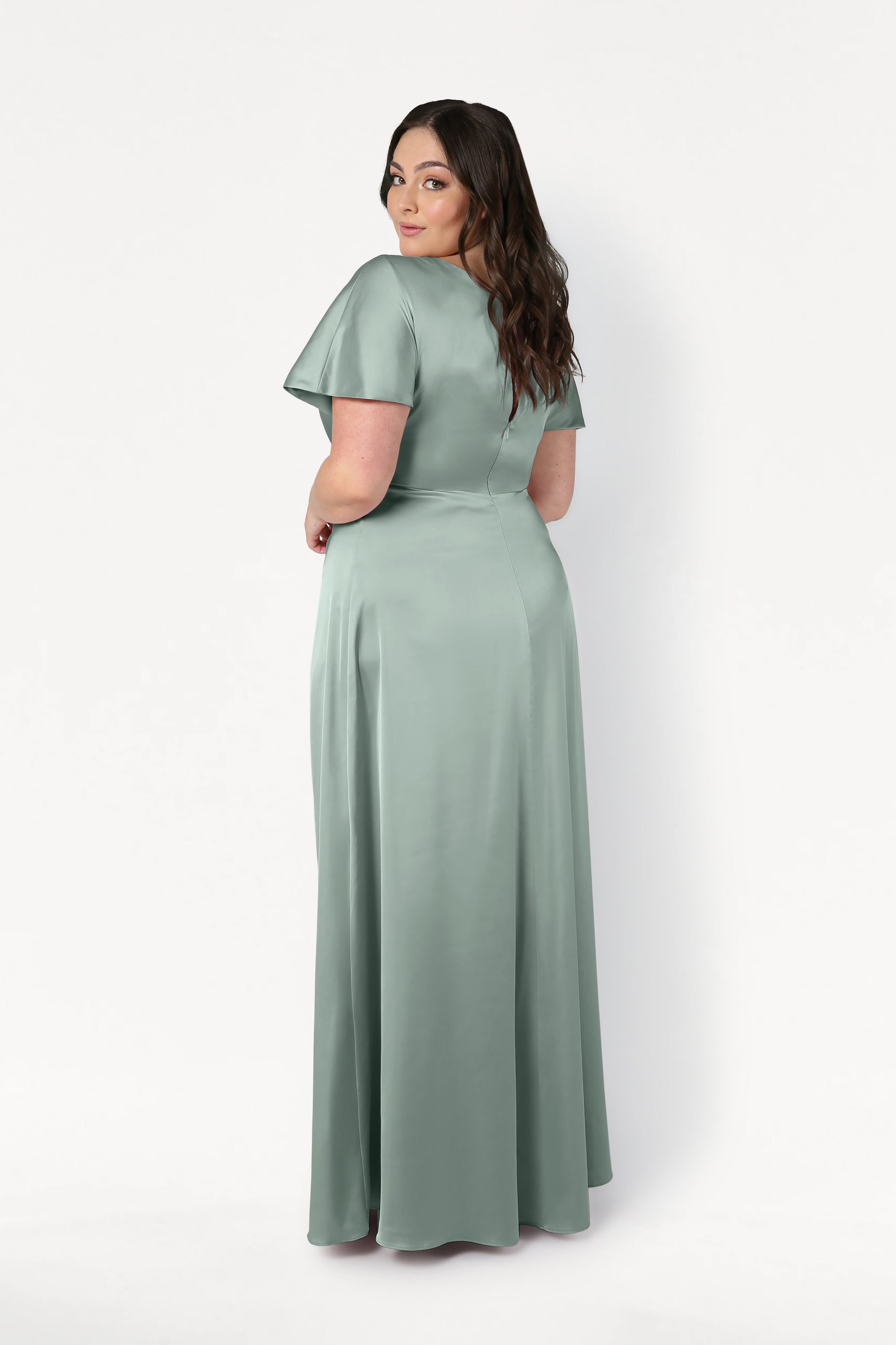 A woman with long brown hair models the TH&TH Camilla Bridesmaid Dress in Dusty Eucalyptus Satin, a short-sleeved, floor-length gown with a twist-knot bodice, standing facing away and glancing over her shoulder against a white background.