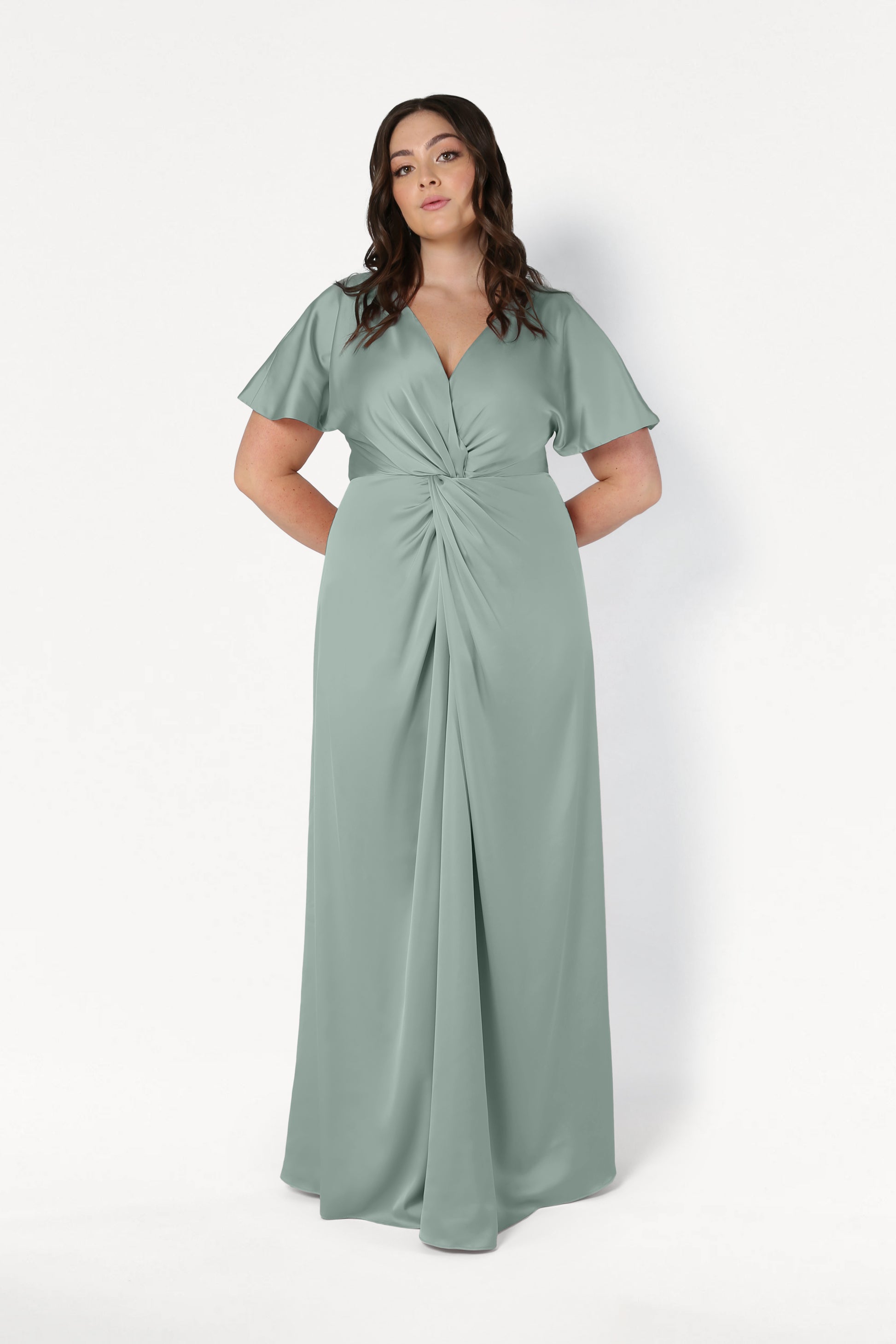 A woman poses against a white background in the TH&TH Camilla Bridesmaid Dress in Dusty Eucalyptus Satin, featuring short flutter sleeves, a V-neckline, and an elegant twist-knot bodice. Her hands rest behind her back.