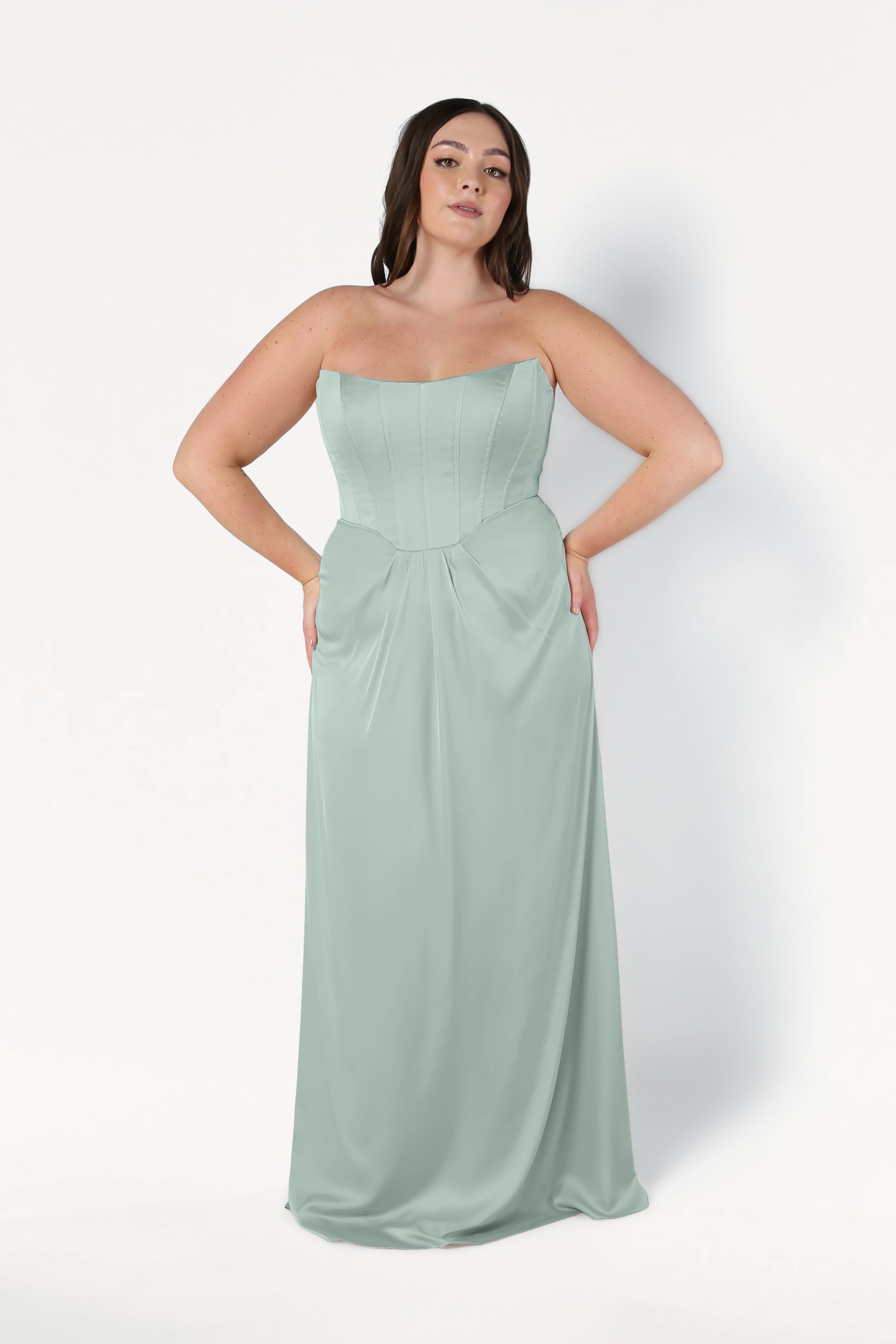 A woman with long, dark hair stands against a white background in the TH&TH Selena Bridesmaid Dress in Dusty Eucalyptus Satin, a size-inclusive strapless gown with a fitted corset bodice and flowing floor-length skirt.